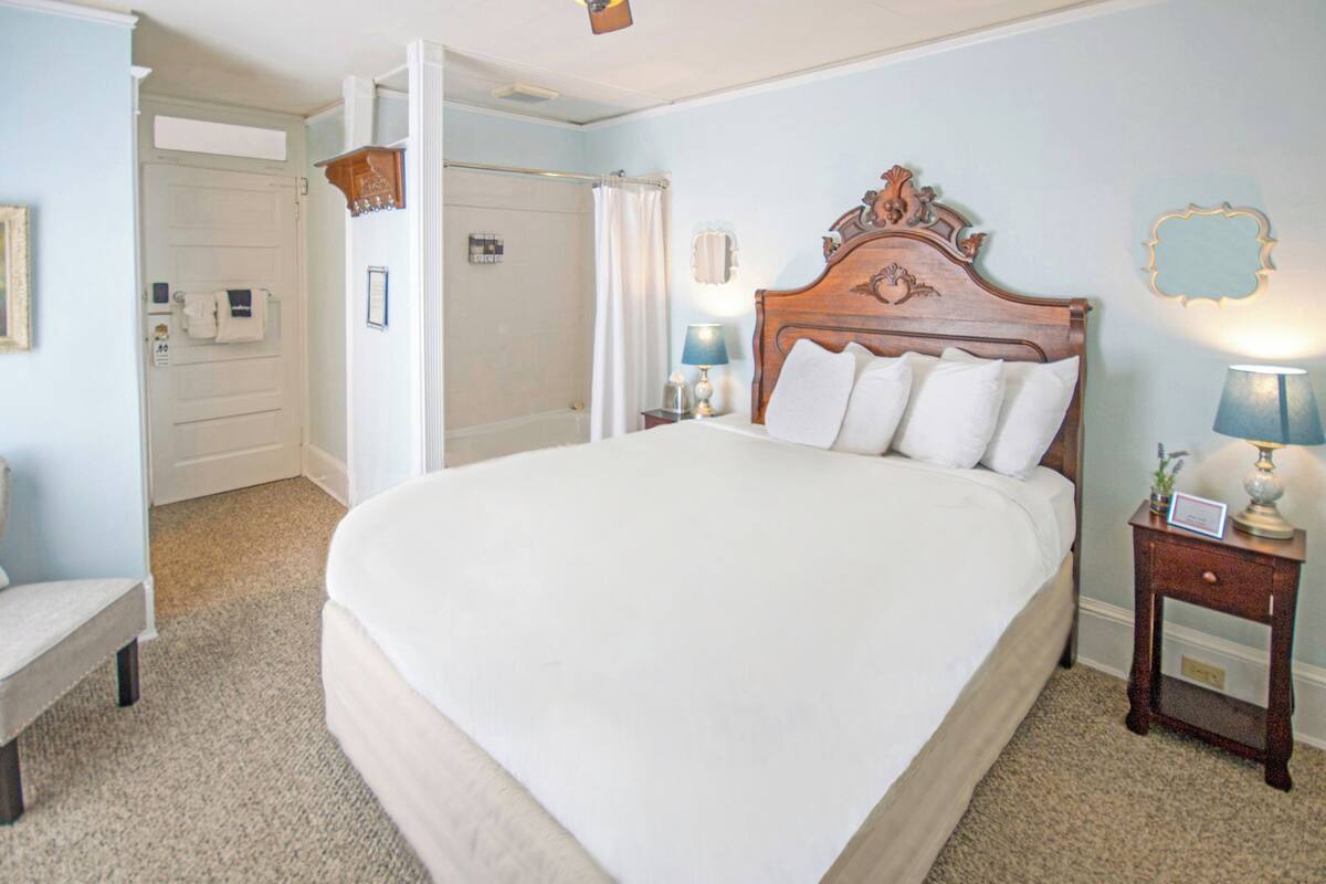 A large bed with a wooden headboard, two bedside tables with lamps, a chair, and a door leading to a bathroom.