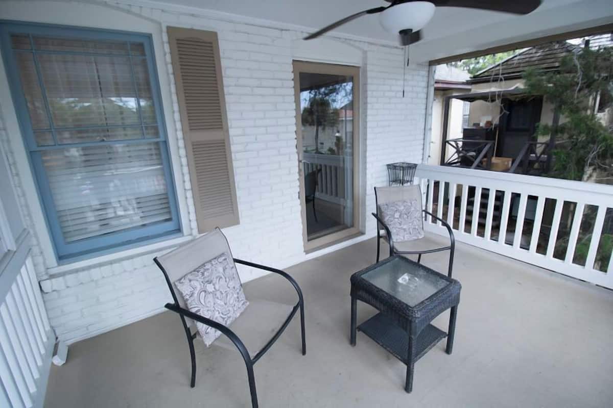 A covered porch with two chairs and a table, white railings, and a door leading to an outdoor area.