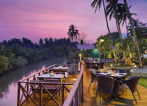 An outdoor dining area with tables and chairs, overlooking a river and surrounded by palm trees. - Lazy Lagoon, Baga A Lemon Tree Resort (Arpora)