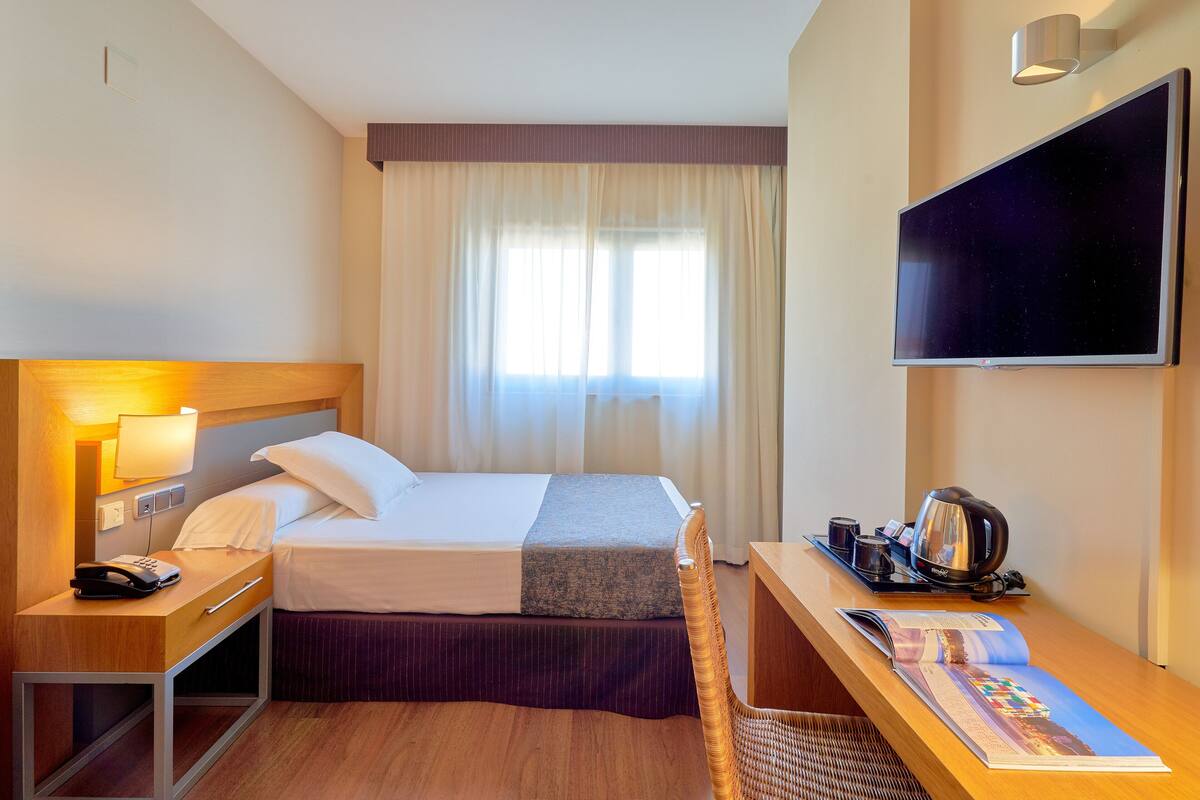 Habitación de hotel con cama, mesita de noche con lámpara, televisor montado en la pared, ventana con cortinas y una revista sobre la mesa.