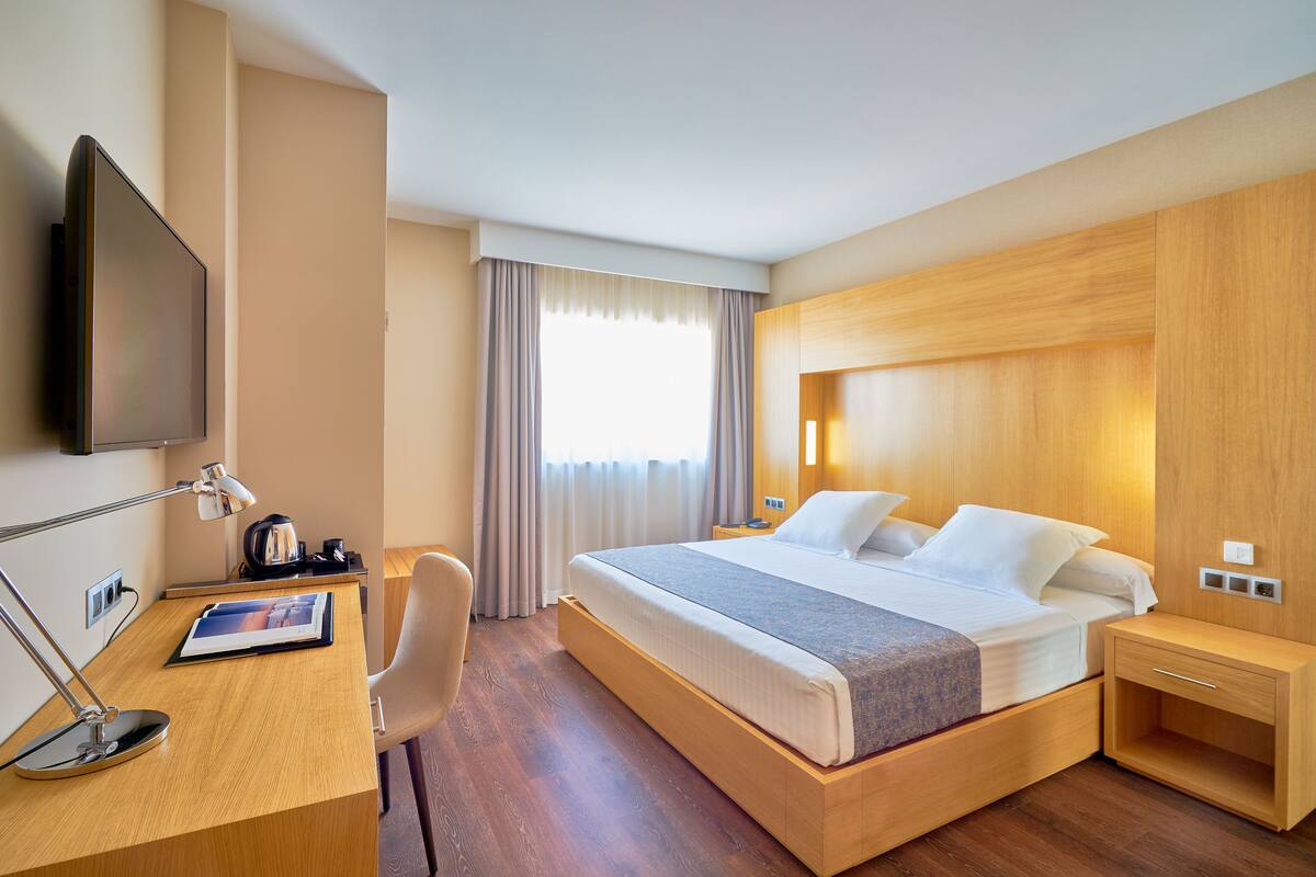 Habitación de hotel con una cama grande, un escritorio con silla, un televisor de pantalla plana y una ventana con cortinas.
