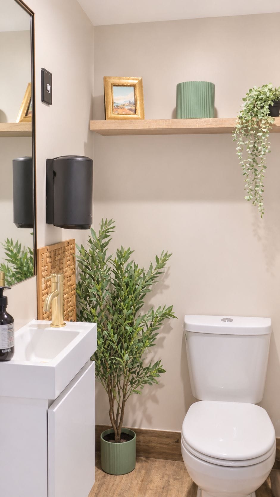 Un baño con lavamanos blanco, inodoro, espejo, planta verde y un cuadro en un estante.