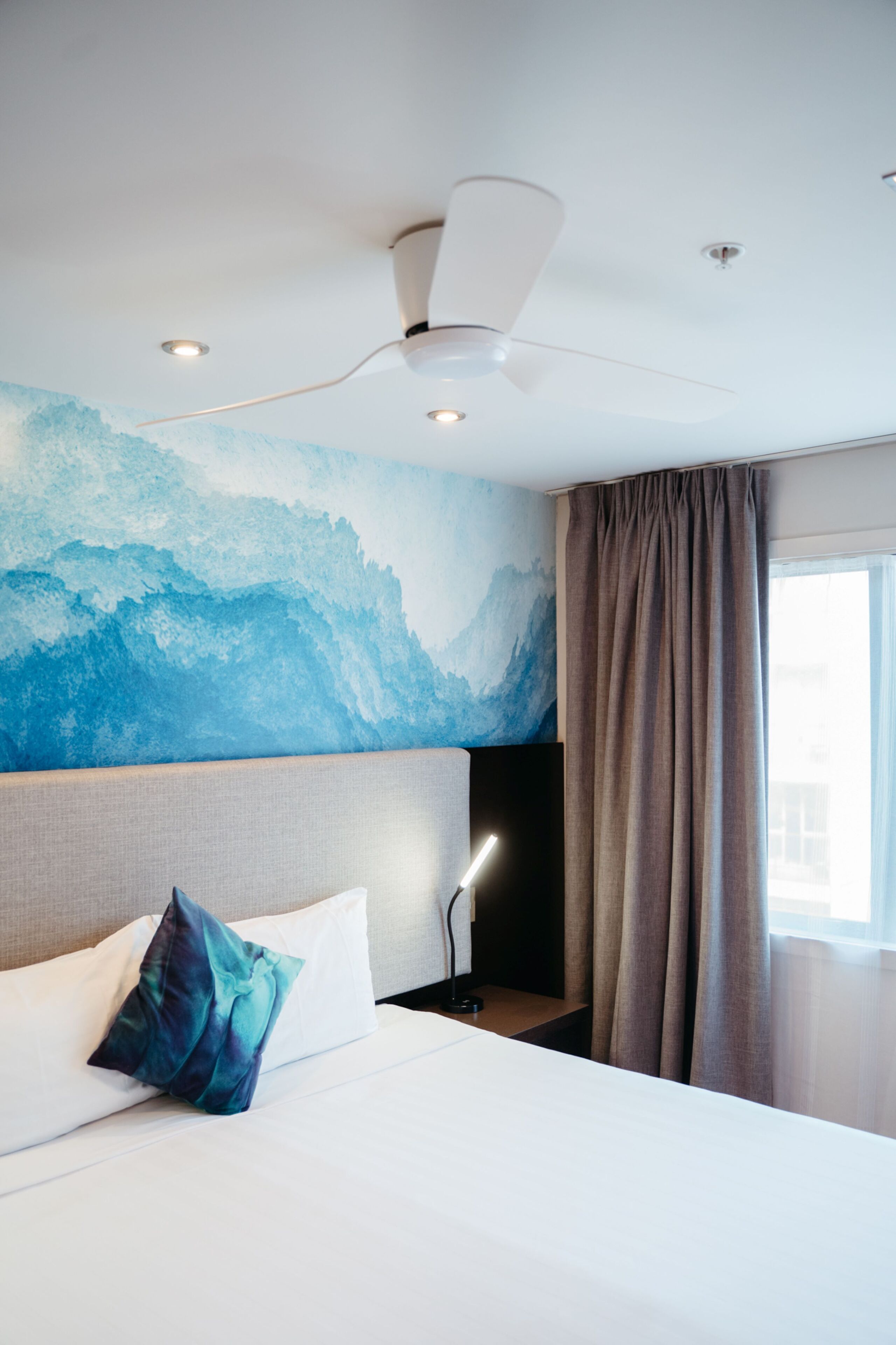 A modern hotel room with a large bed, a ceiling fan, recessed lighting, a mountain mural, and a view of the city through the window.