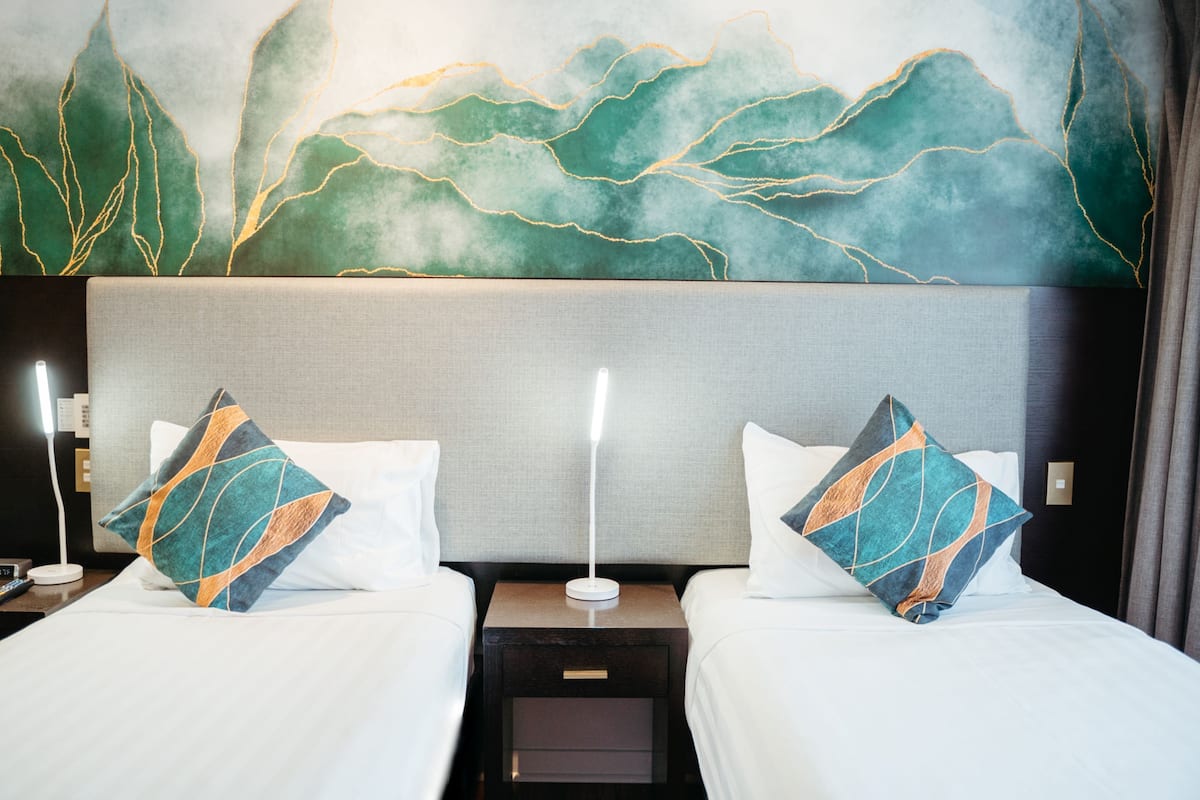 Une chambre d’hôtel avec deux lits, chacun recouvert d’une couvre-lit blanche et agrémenté d’un coussin décoratif. Une table de chevet se trouve entre les lits, avec une lampe et une prise électrique. Le mur derrière les lits est orné d’un grand tableau abstrait aux tons verts et dorés.