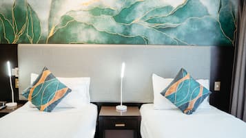 Habitación de hotel con dos camas, cada una con una colcha blanca y un cojín decorativo. Una mesita de noche entre las camas tiene una lámpara y un enchufe. La pared detrás de las camas presenta un cuadro abstracto de tonos verdes y dorados.
