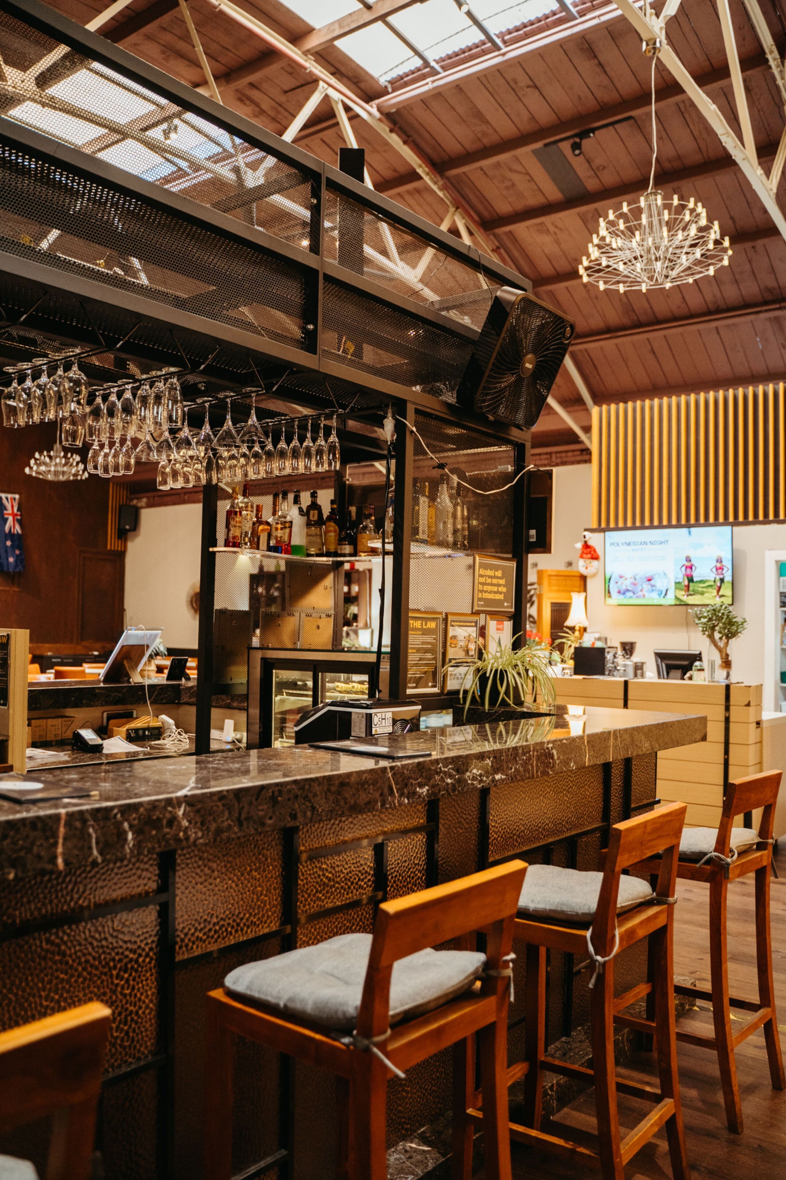 A bar area with a long counter, multiple stools, hanging glasses, and a television screen displaying a sports game.