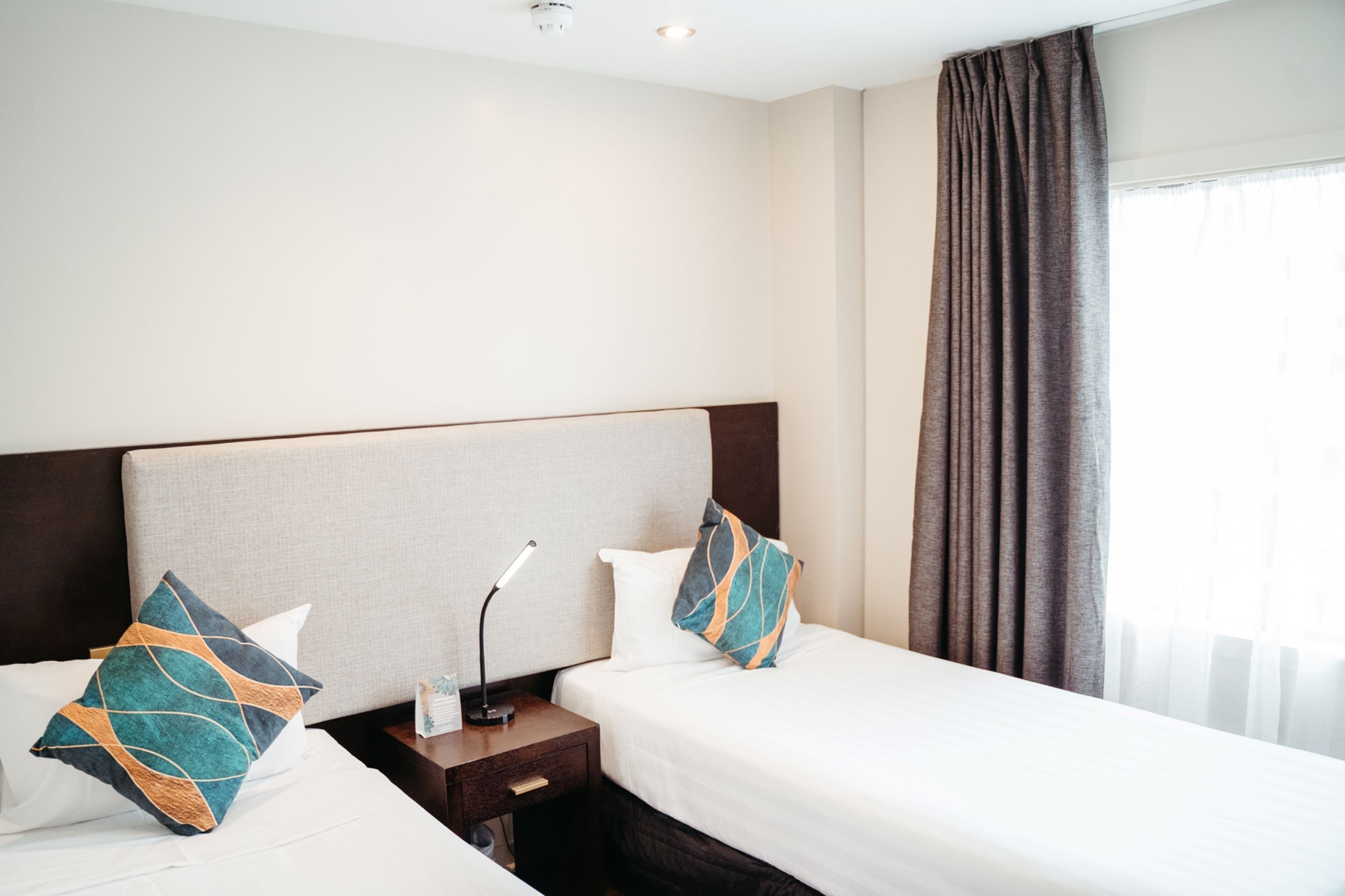 Habitación de hotel con dos camas, una mesita de noche con lámpara y un ventanal con cortinas.