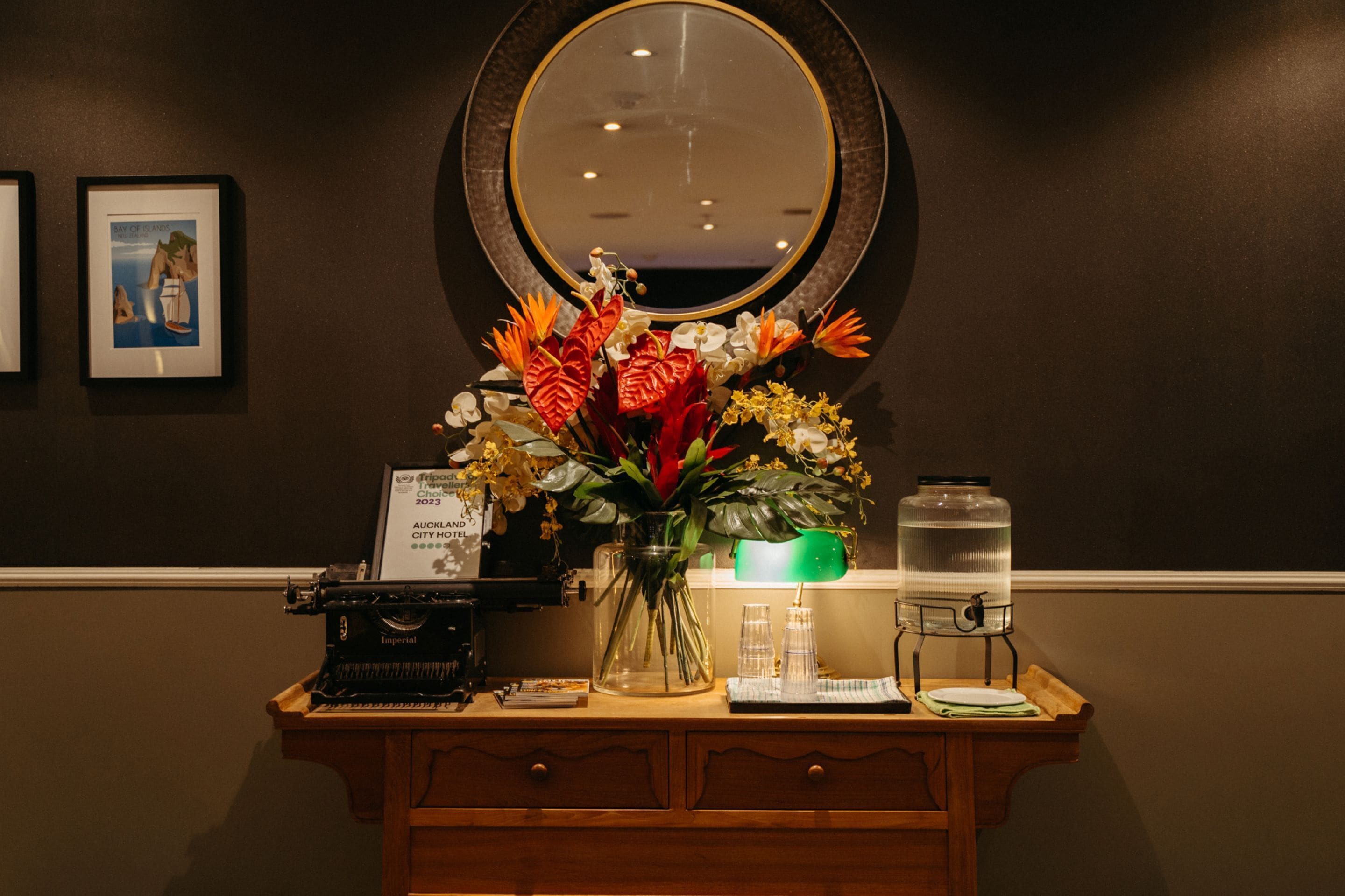 A reception desk with a mirror, framed artwork, a vase of flowers, a water dispenser, and a vintage typewriter.