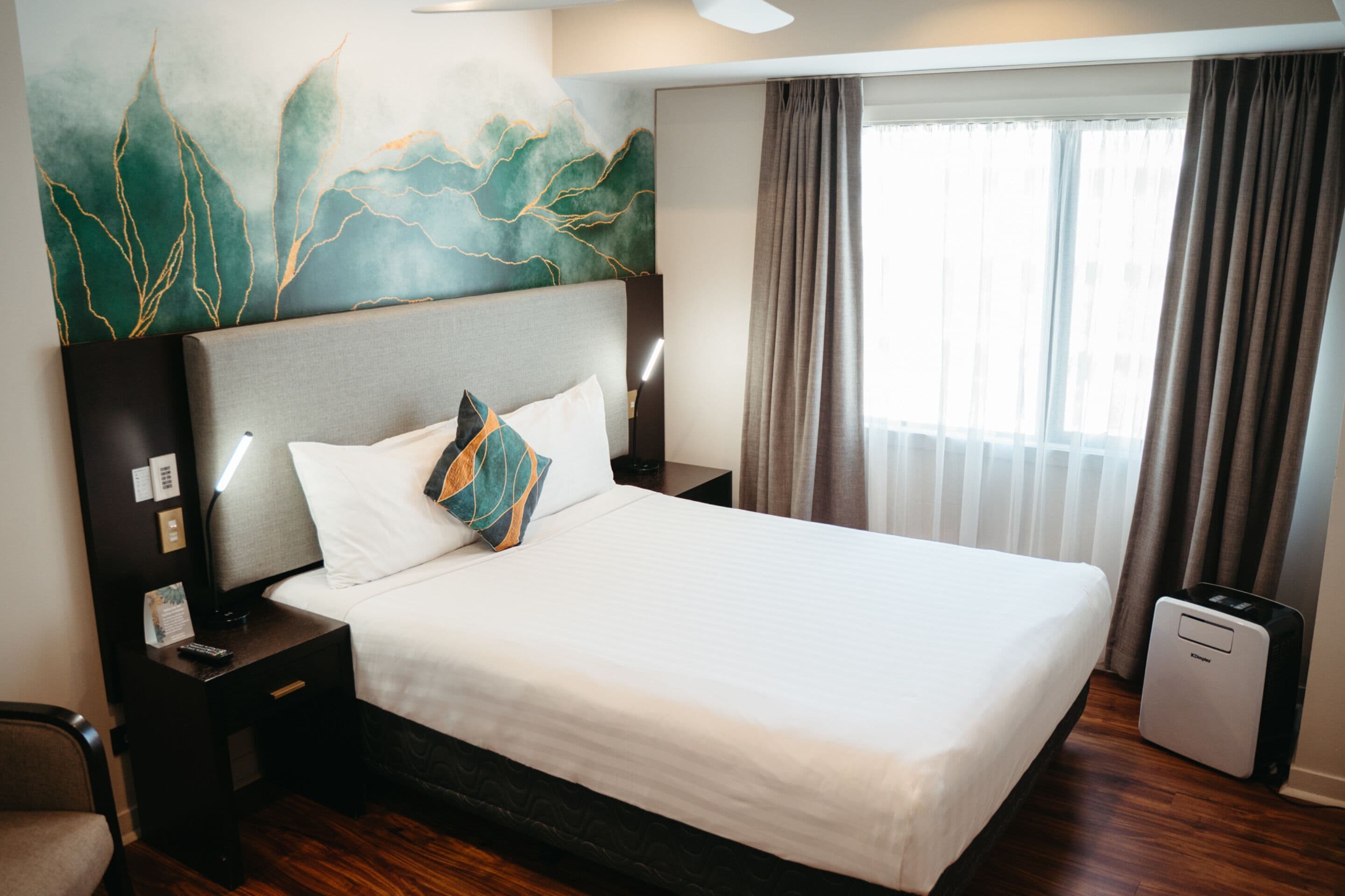 A hotel room with a large bed, a nightstand, a lamp, a wall art piece, a window with curtains, and a small heater.