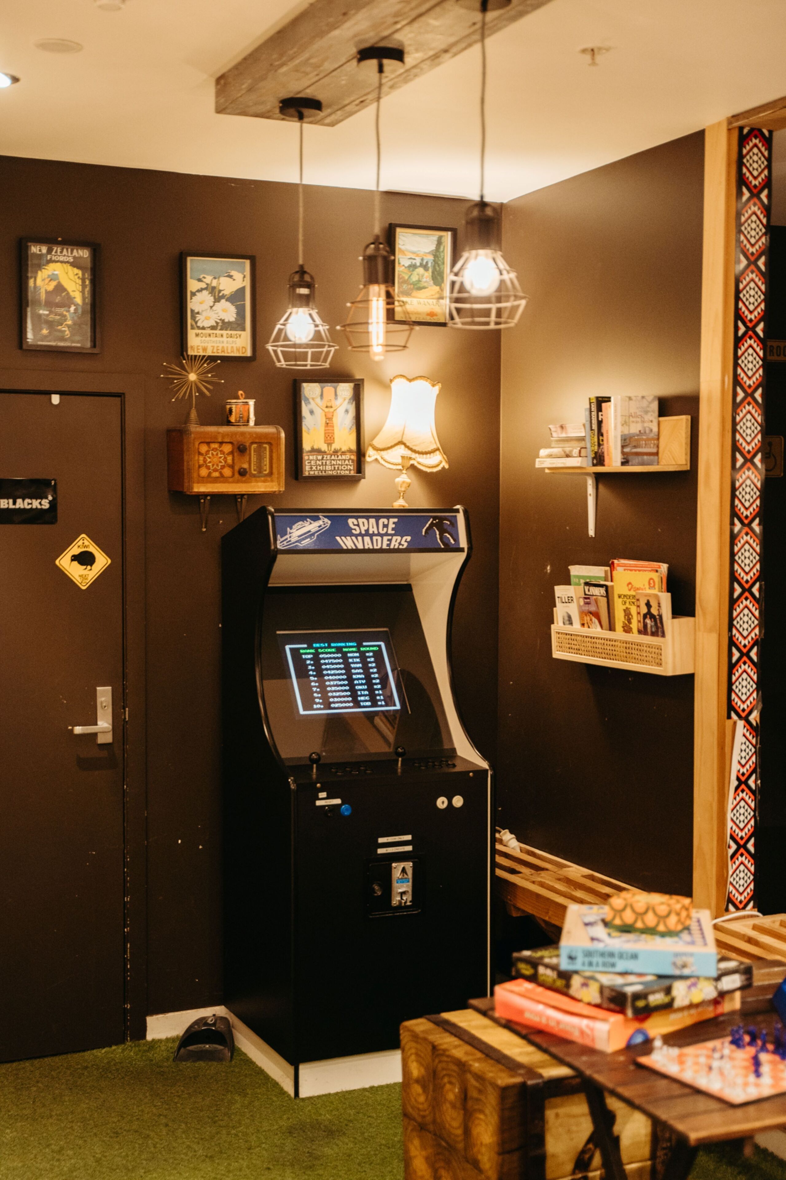 Ein Zimmer mit einem Spielautomaten zum Thema Weltraum, einem Holztisch mit Büchern und einer Wand mit gerahmten Bildern sowie einer Tür mit der Aufschrift 'BLACKS'.