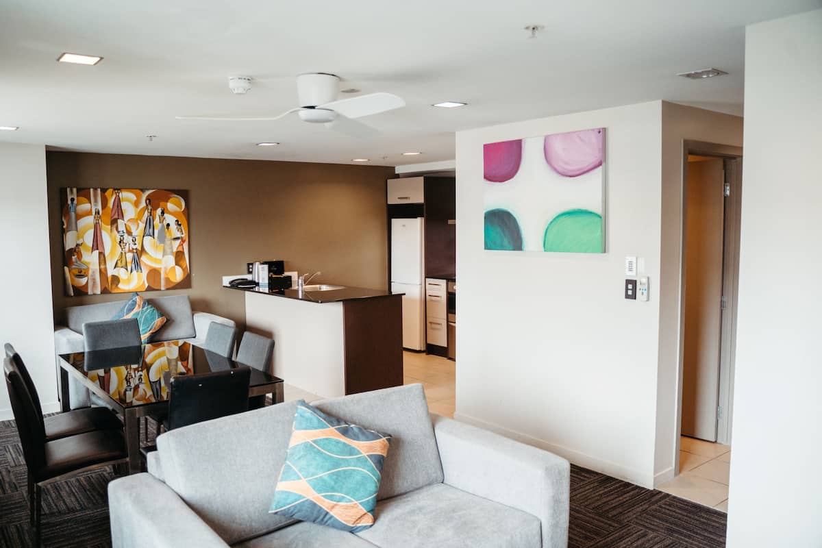 Une chambre d’hôtel moderne dotée d’un coin repas, d’une kitchenette et d’un espace de vie avec un canapé gris et des œuvres d’art colorées.