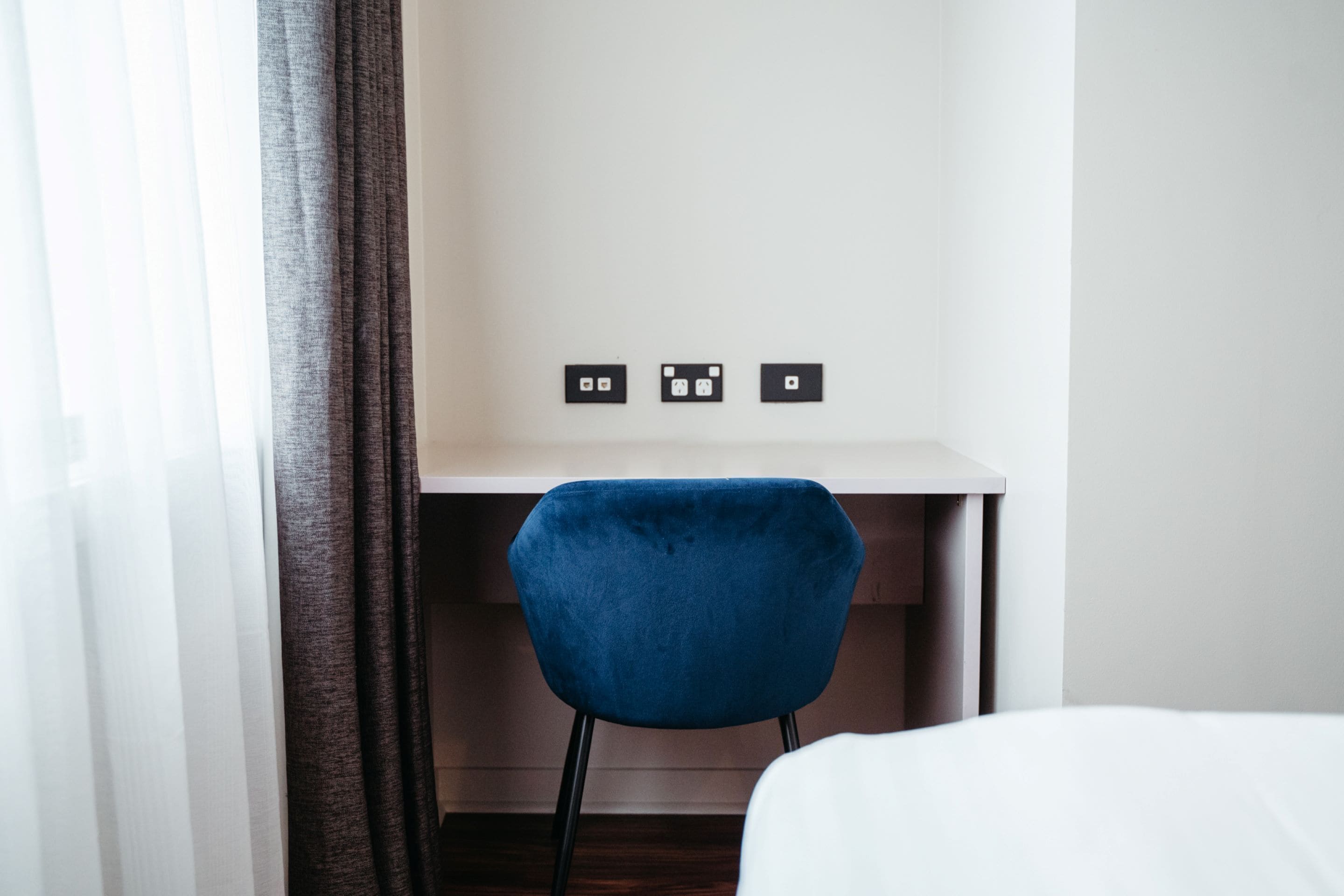 A hotel room with a bed, a desk with a chair, and three wall sockets.