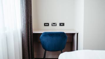 A hotel room with a bed, a desk with a chair, and three wall sockets.