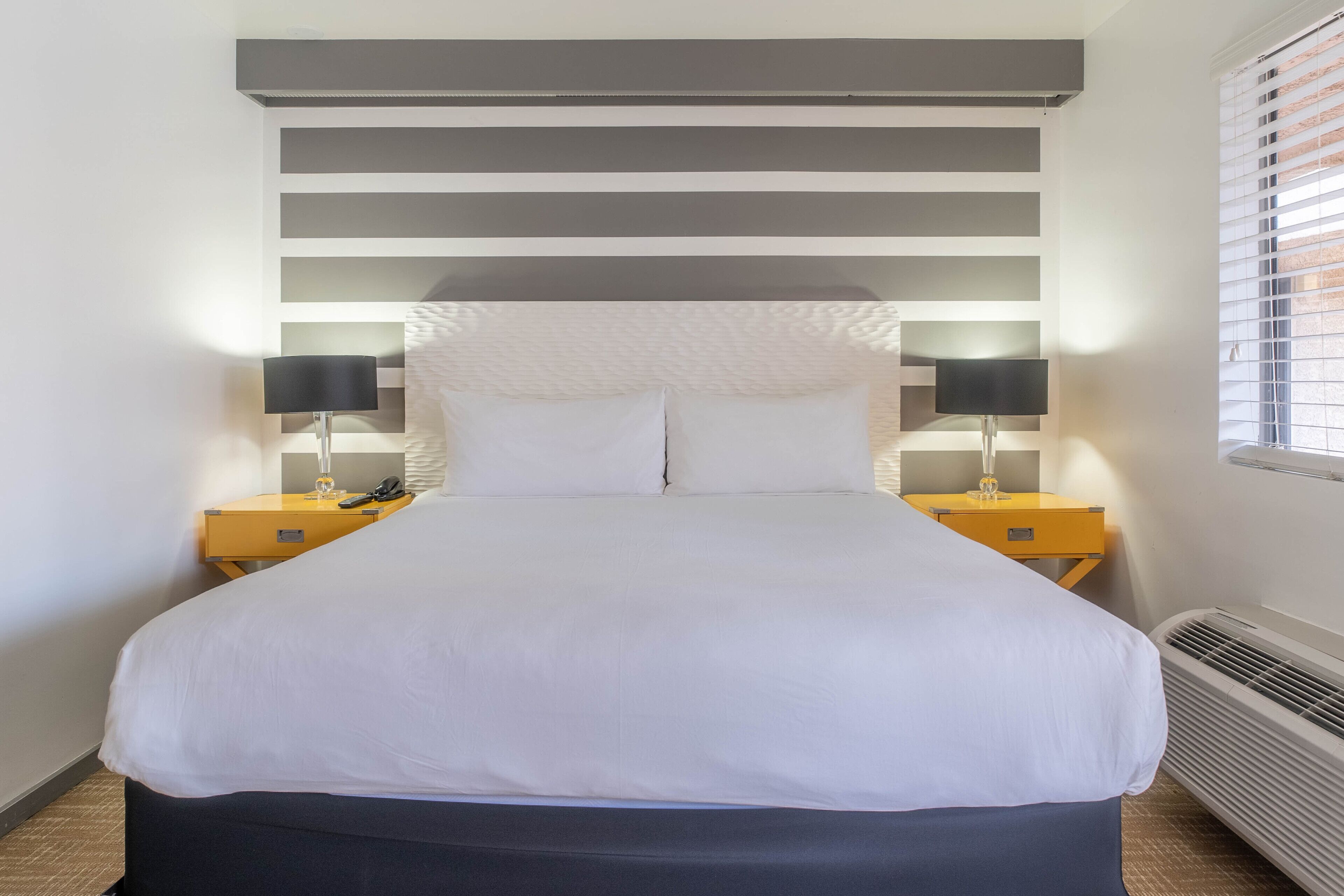 A neatly made bed with white linens, two bedside tables with lamps, and a window with blinds.