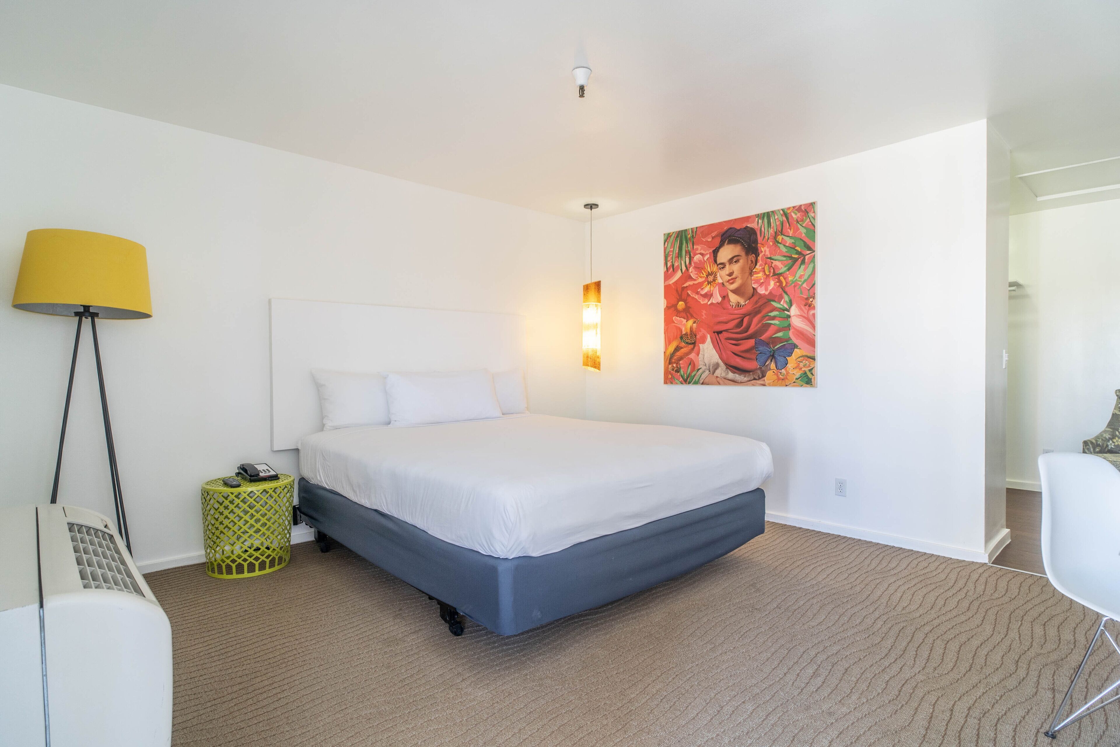 A modern hotel room with a large bed, a standing lamp, a small side table, and a colorful painting on the wall.