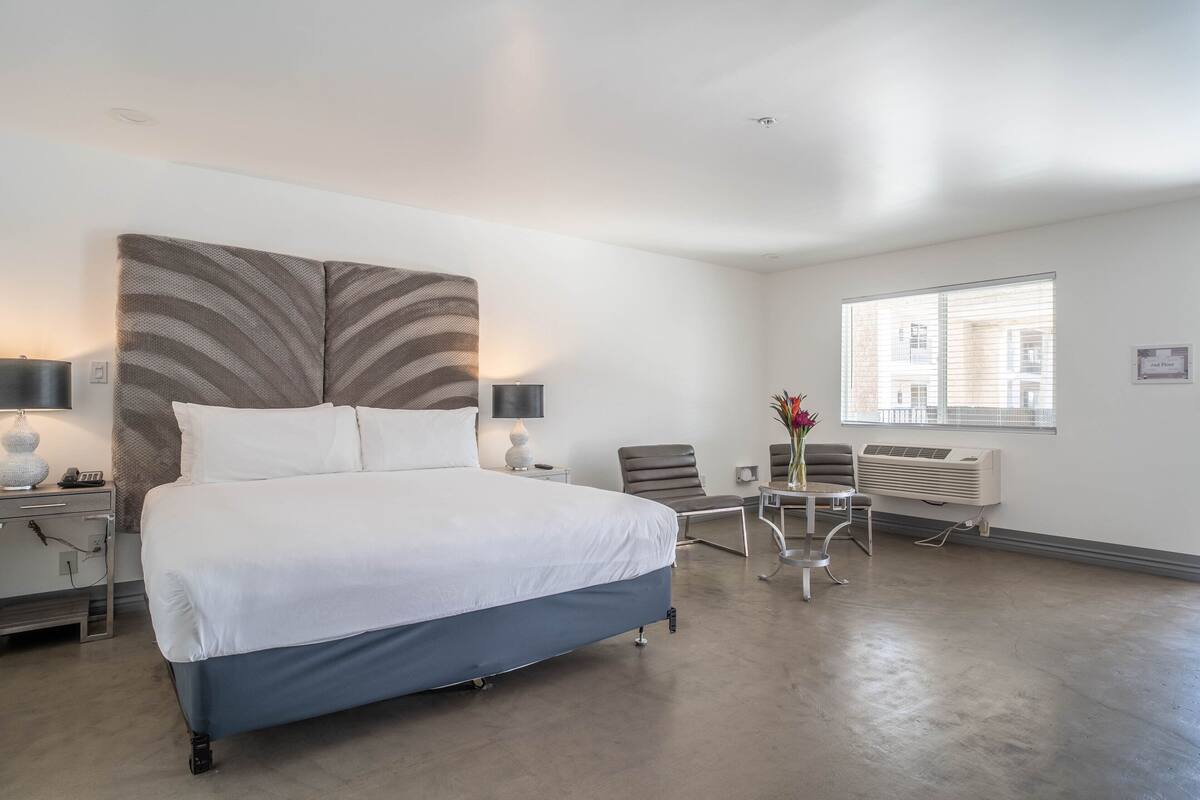 Ein modernes Hotelzimmer mit einem großen Bett, einem Schreibtisch mit Stuhl, einem kleinen Tisch mit Vase und einem Fenster mit Jalousien.