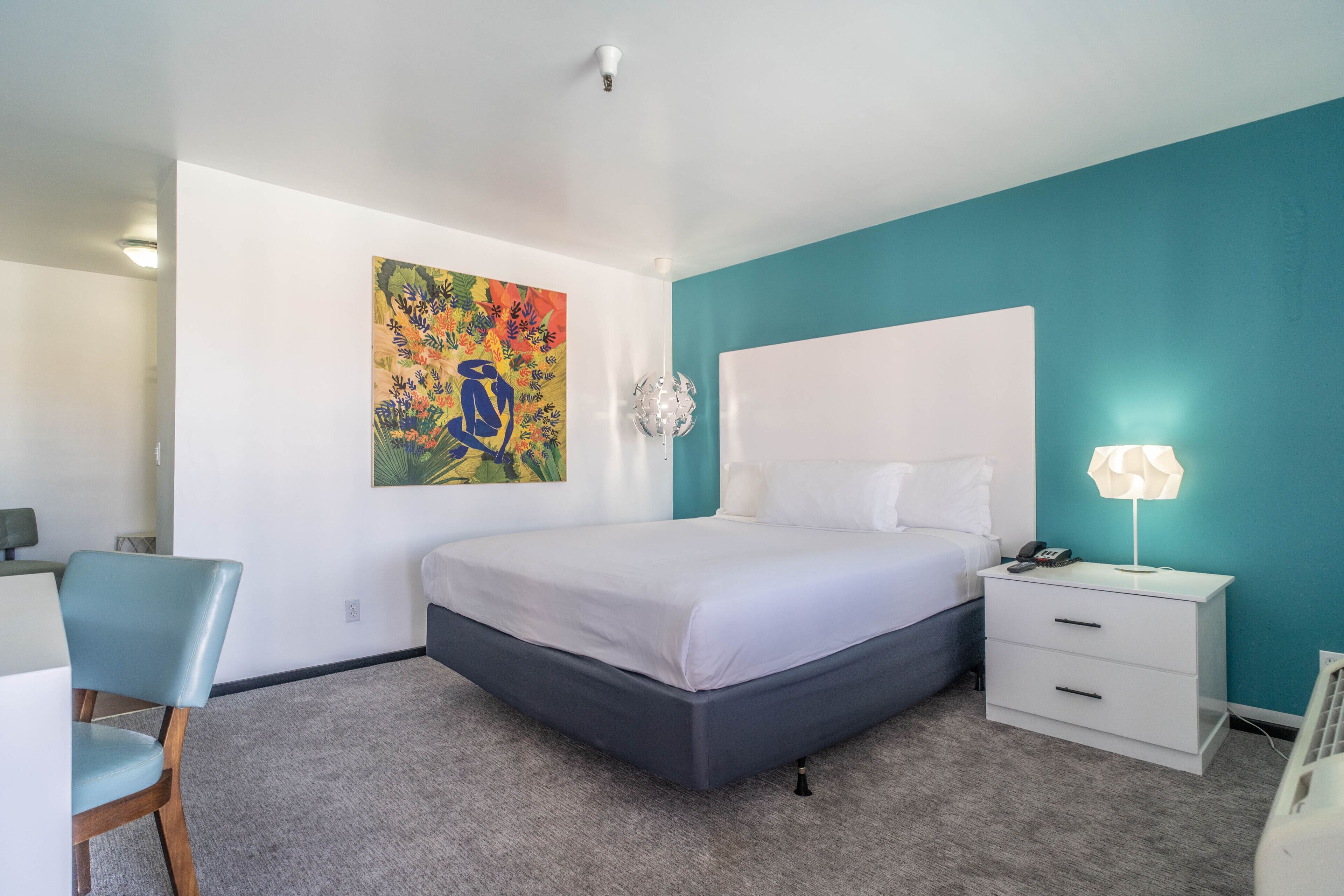 A bedroom with a bed, a desk, a chair, a nightstand, and a wall painting.