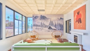 Een kamer met een groene bank, twee rieten stoelen, een open haard en een groot raam met uitzicht op palmbomen.