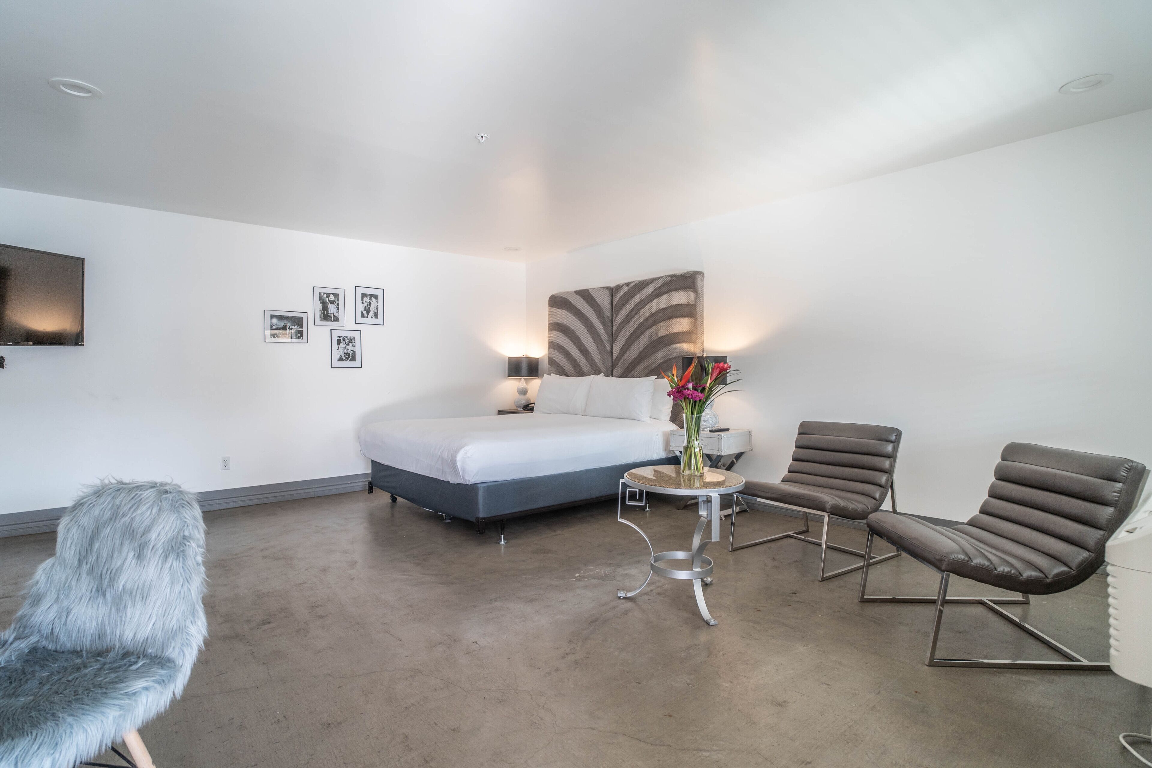 Une chambre d’hôtel moderne avec un grand lit, deux fauteuils, une petite table ronde ornée d’un vase de fleurs et un téléviseur à écran plat fixé au mur.