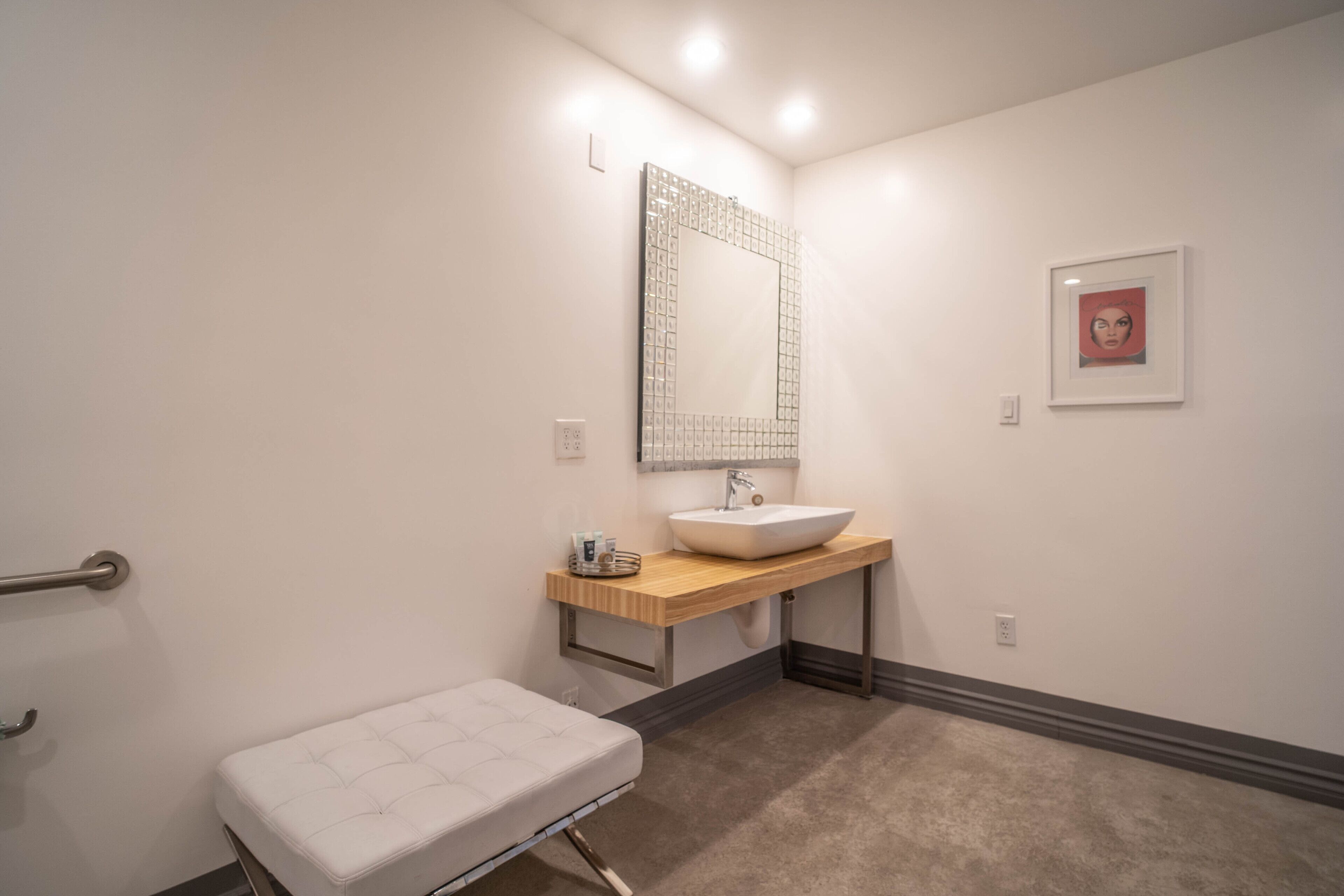 Un baño con un lavamanos blanco, un espejo, un mueble de tocador de madera, un banco blanco y un cuadro enmarcado en la pared.