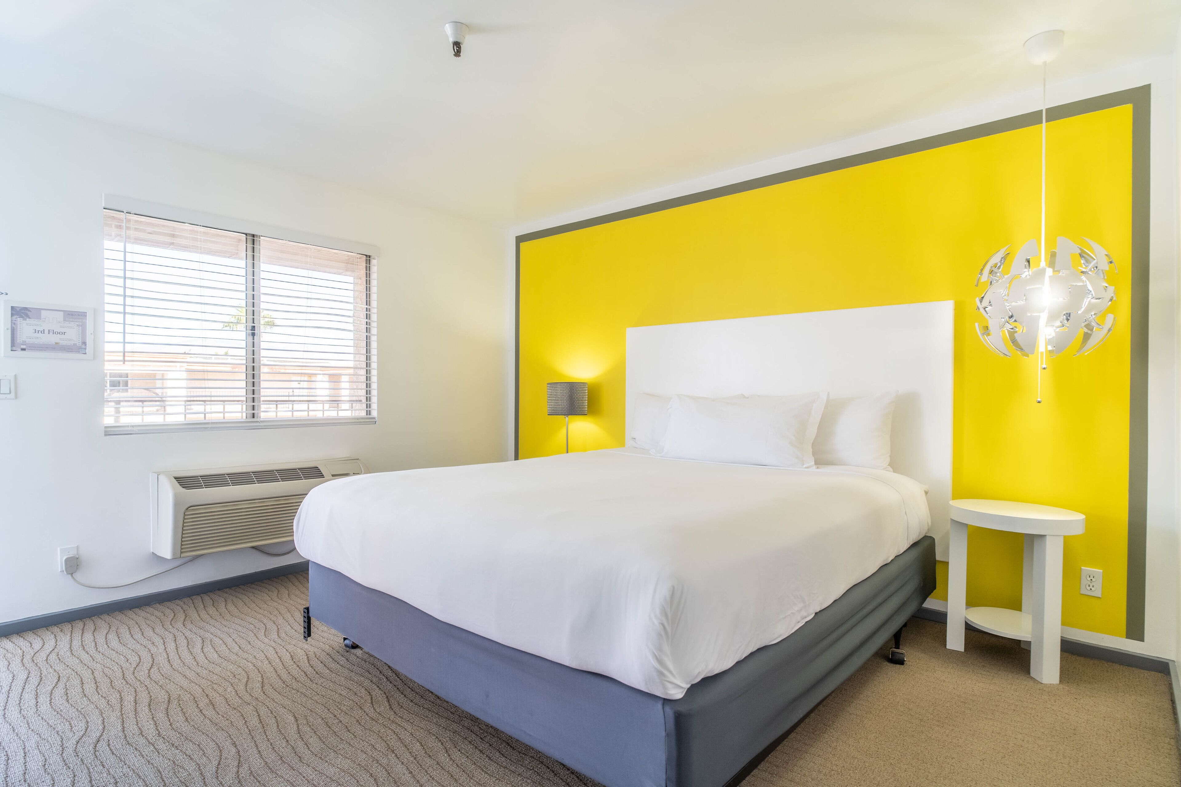 A bedroom with a bed, a bedside table, a lamp, a window with blinds, and a wall-mounted air conditioner.