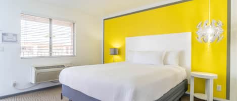 A bedroom with a bed, a bedside table, a lamp, a window with blinds, and a wall-mounted air conditioner.