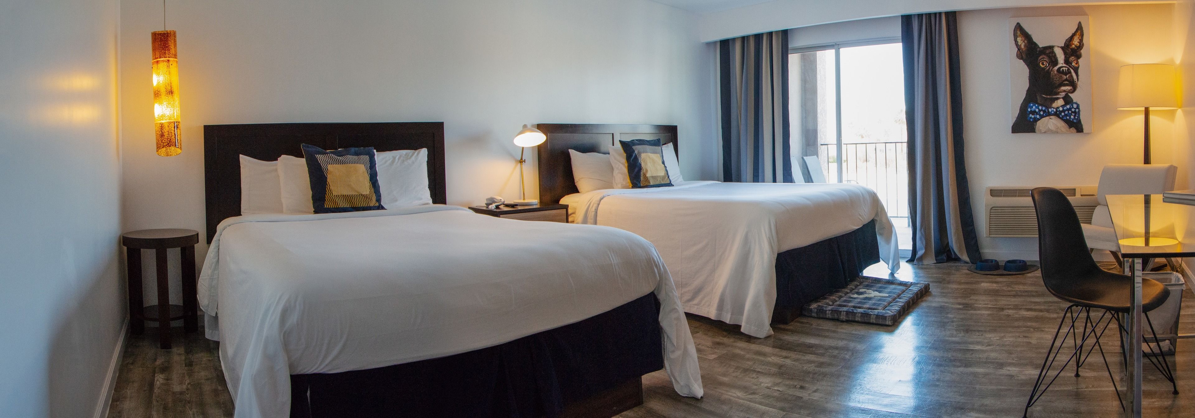 A hotel room with two beds, a desk, a chair, a lamp, and a large window with curtains.