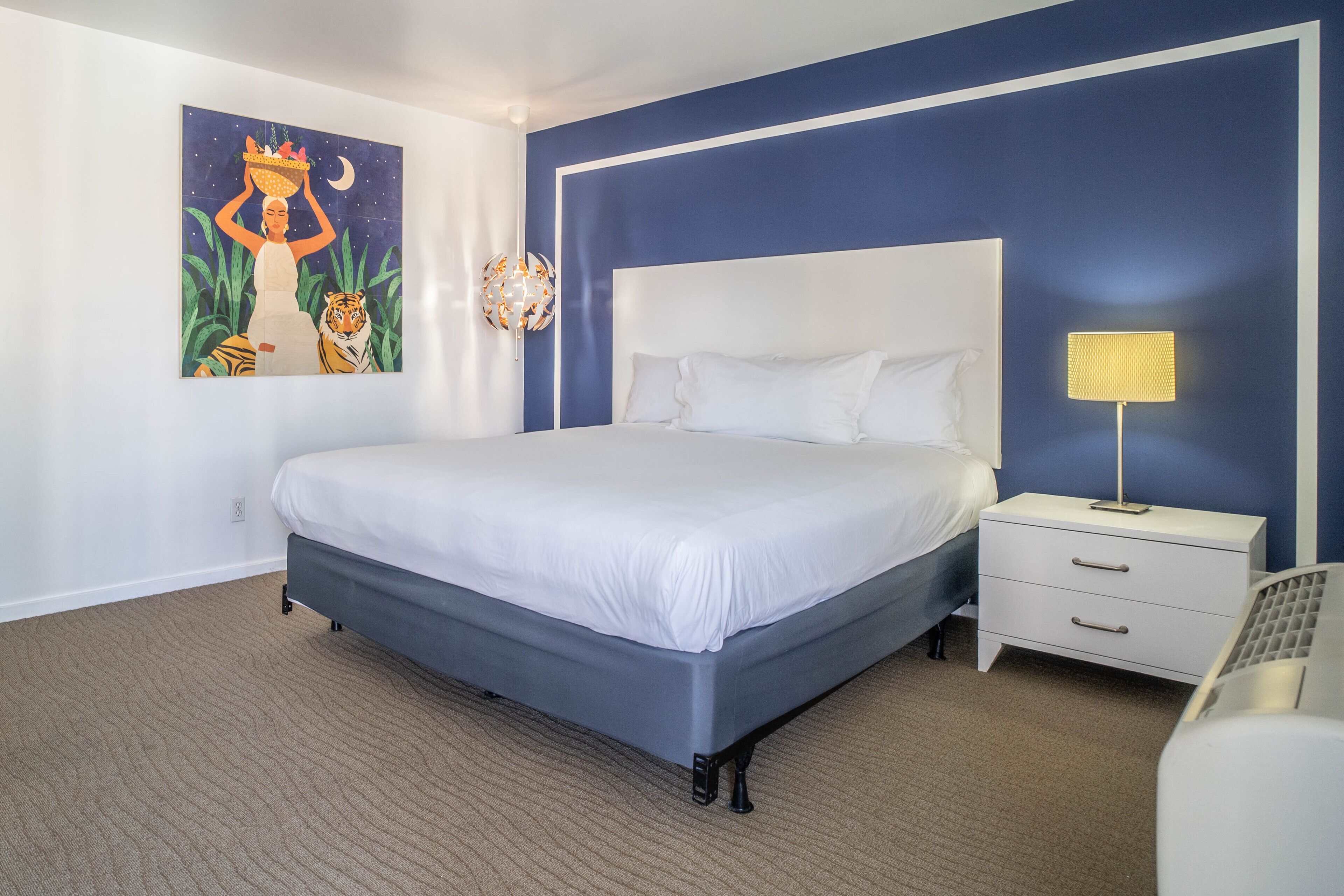 A bedroom with a large bed, a nightstand with a lamp, and a wall-mounted artwork.