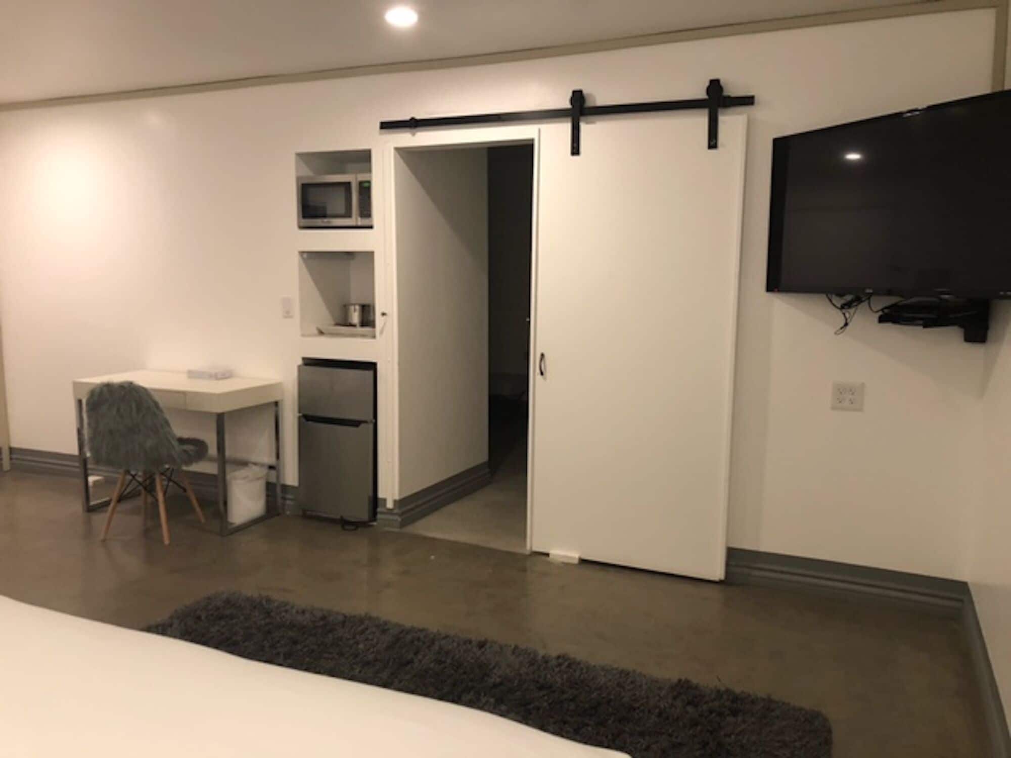 A modern bedroom with a bed, a desk, a chair, a wardrobe, a television, and a small kitchenette.