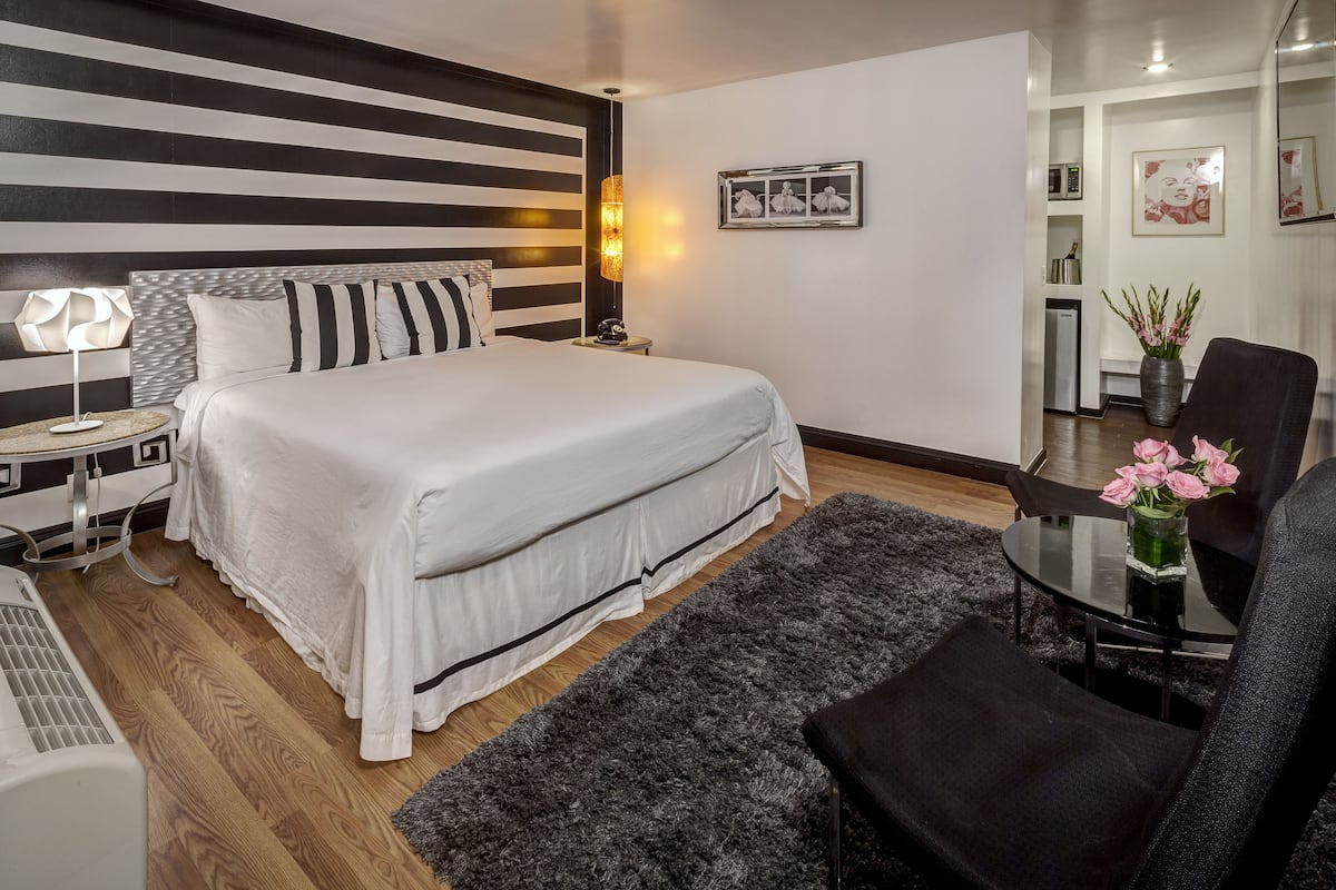 Ein modernes Hotelzimmer mit einem großen Bett, einem Nachttisch mit Lampe, einem kleinen runden Tisch mit Blumenvase und einem Sessel.