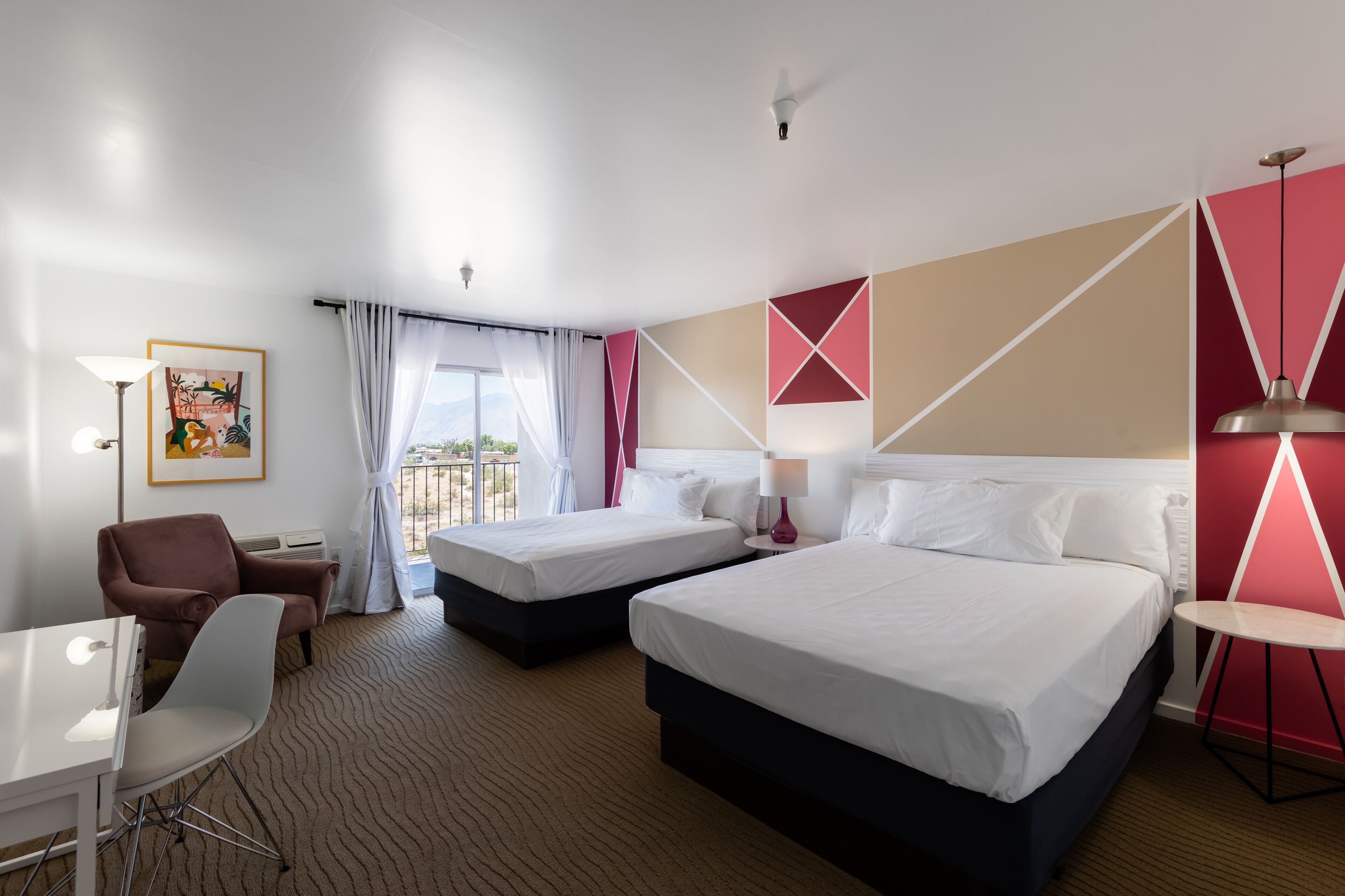 A hotel room with two beds, a desk, a chair, and a view of the outdoors.