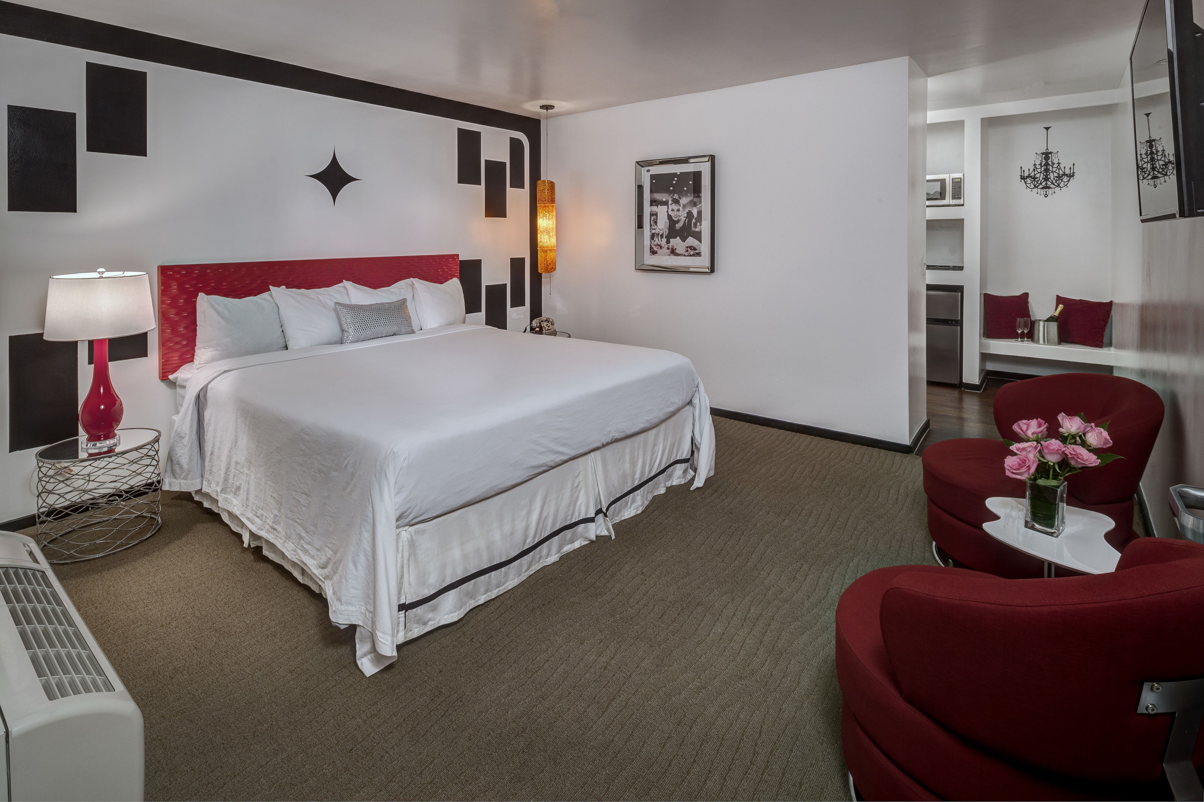 A hotel room with a large bed, a red chair, a small table with flowers, a red lamp, and a picture on the wall.