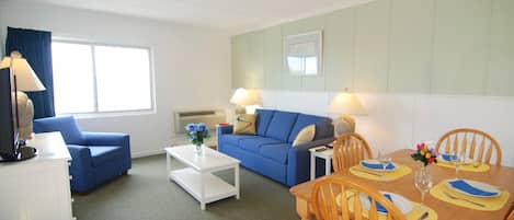 A room with a blue sofa, a white coffee table, a dining table with chairs, and a TV on a stand.