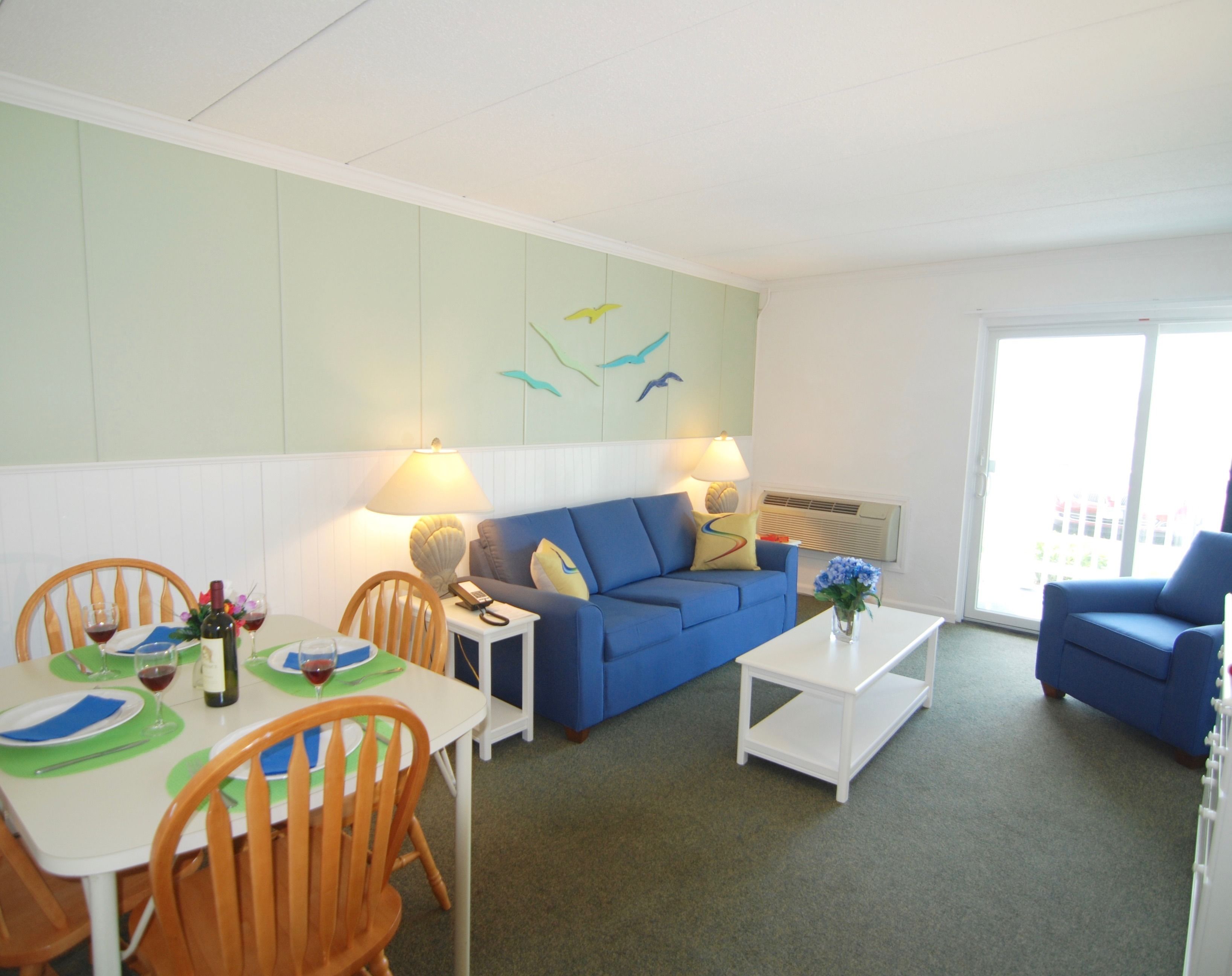 A living room with a blue sofa, a white coffee table, a dining table with chairs, and a wall with a bird mural.