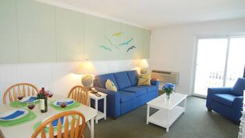 A living room with a blue sofa, a white coffee table, a dining table with chairs, and a wall with a bird mural.