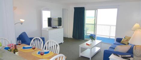 A room with a dining table, chairs, a sofa, a TV, and a balcony with a view.
