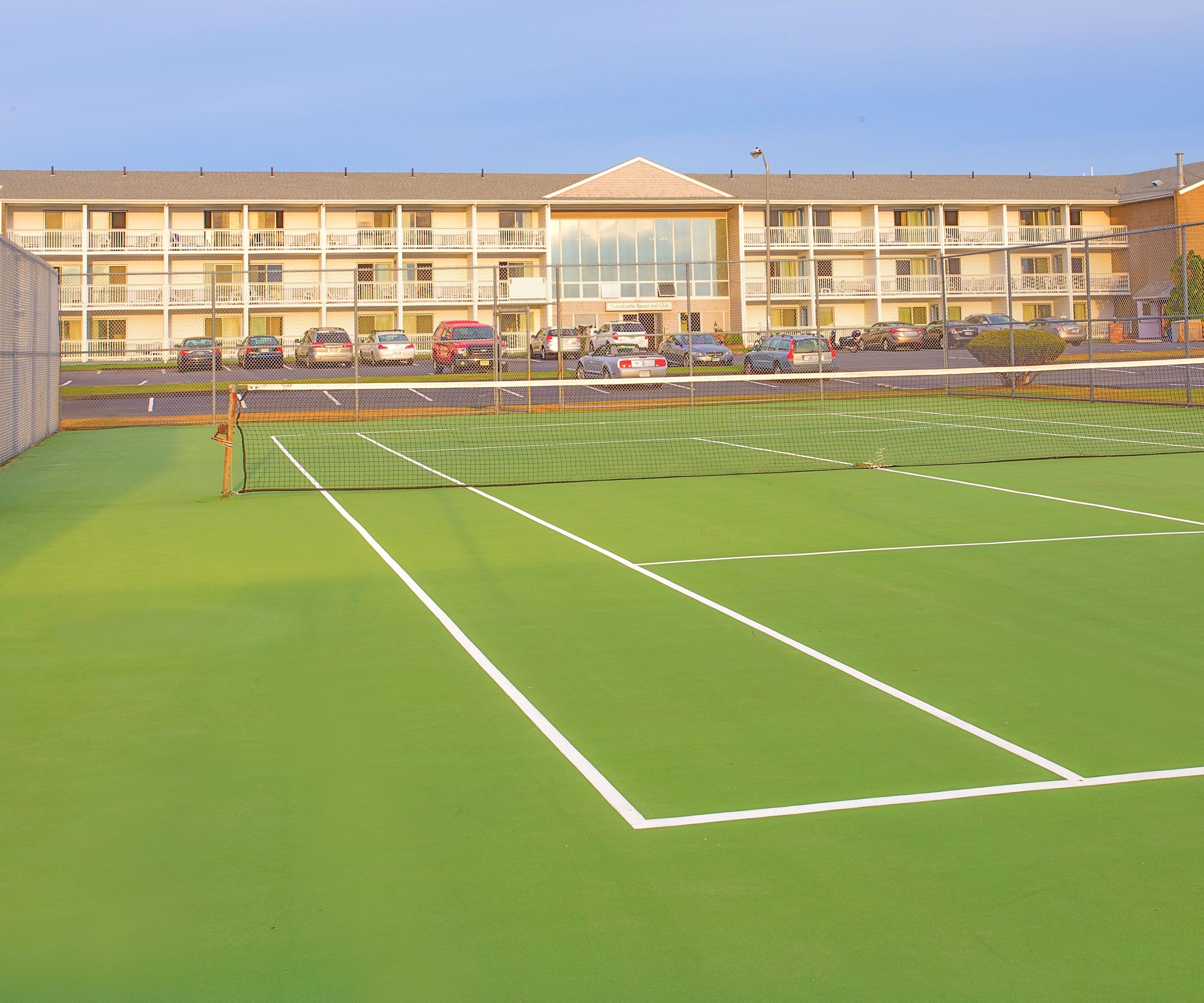 A tennis court with a net, surrounded by a fence, and a building with multiple balconies in the background.