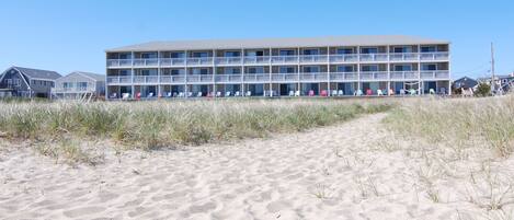 A multi-story hotel with a beachside location, featuring a long row of balconies and a sandy walkway leading to the building.