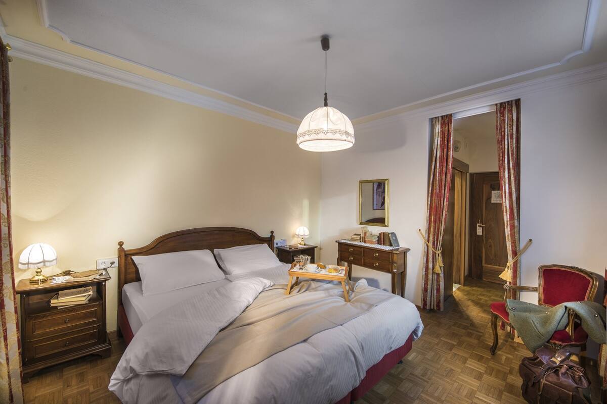 Una cama bien hecha con almohadas y una bandeja con alimentos para el desayuno, una mesita de noche de madera con una lámpara, una silla y un espejo en la pared.