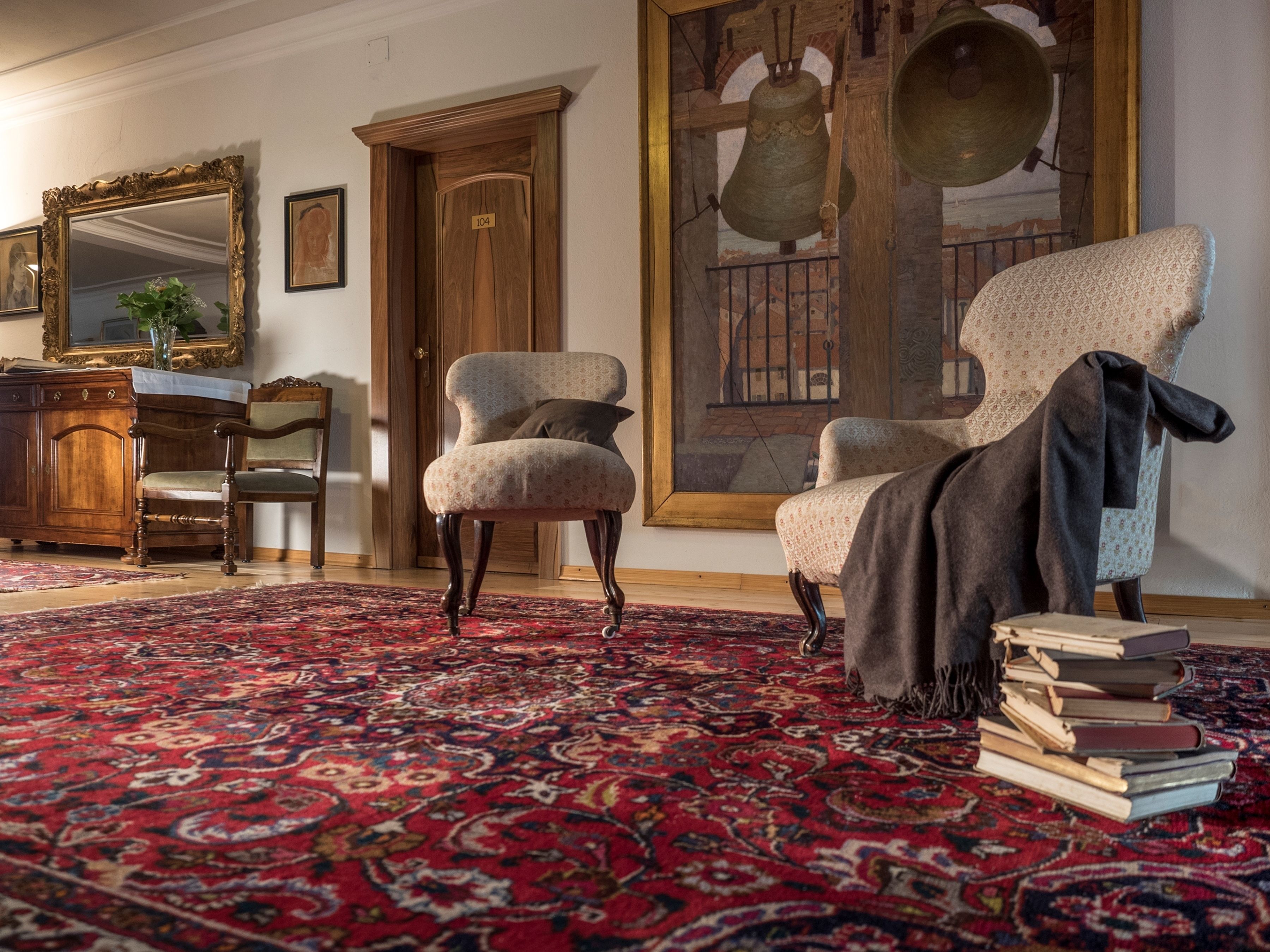 Una habitación con una alfombra grande de estampado, una silla de madera, un escritorio de madera, un espejo y una campana.