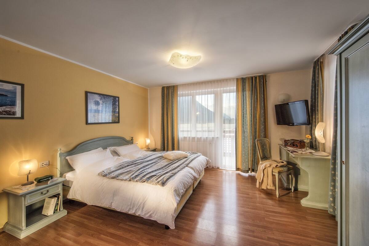 Una habitación de hotel con cama, mesitas de noche, escritorio con silla, televisor y ventana con cortinas.
