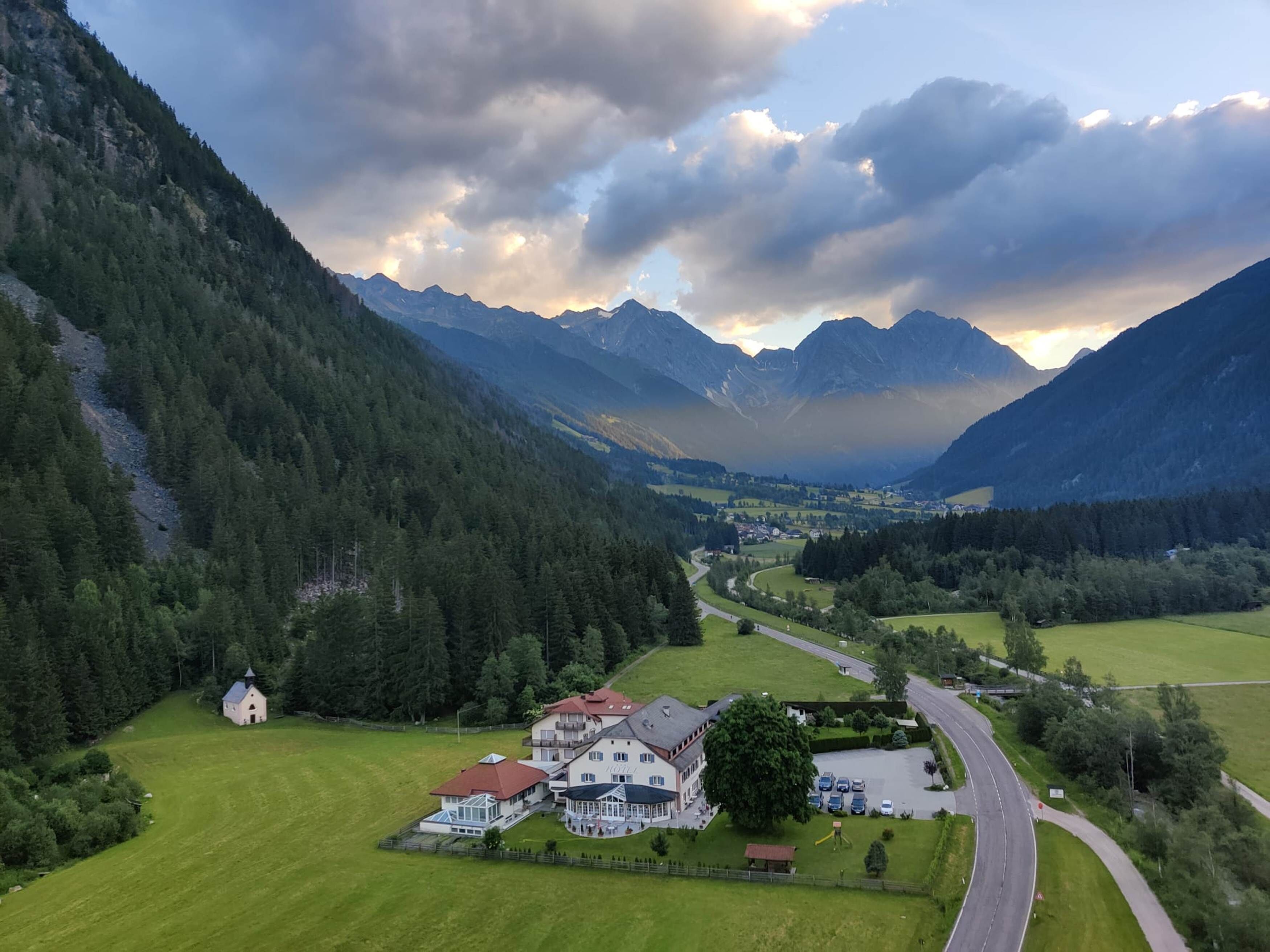 A hotel with a large building and a smaller annex, surrounded by lush green fields and a winding road, with mountains in the background.