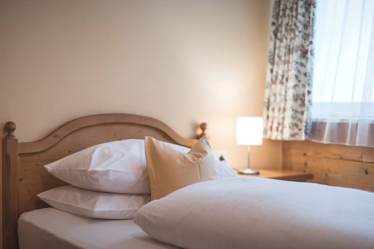 Una cama bien hecha con sábanas y almohadas blancas, un cabecero de madera y una mesita de noche con una lámpara.
