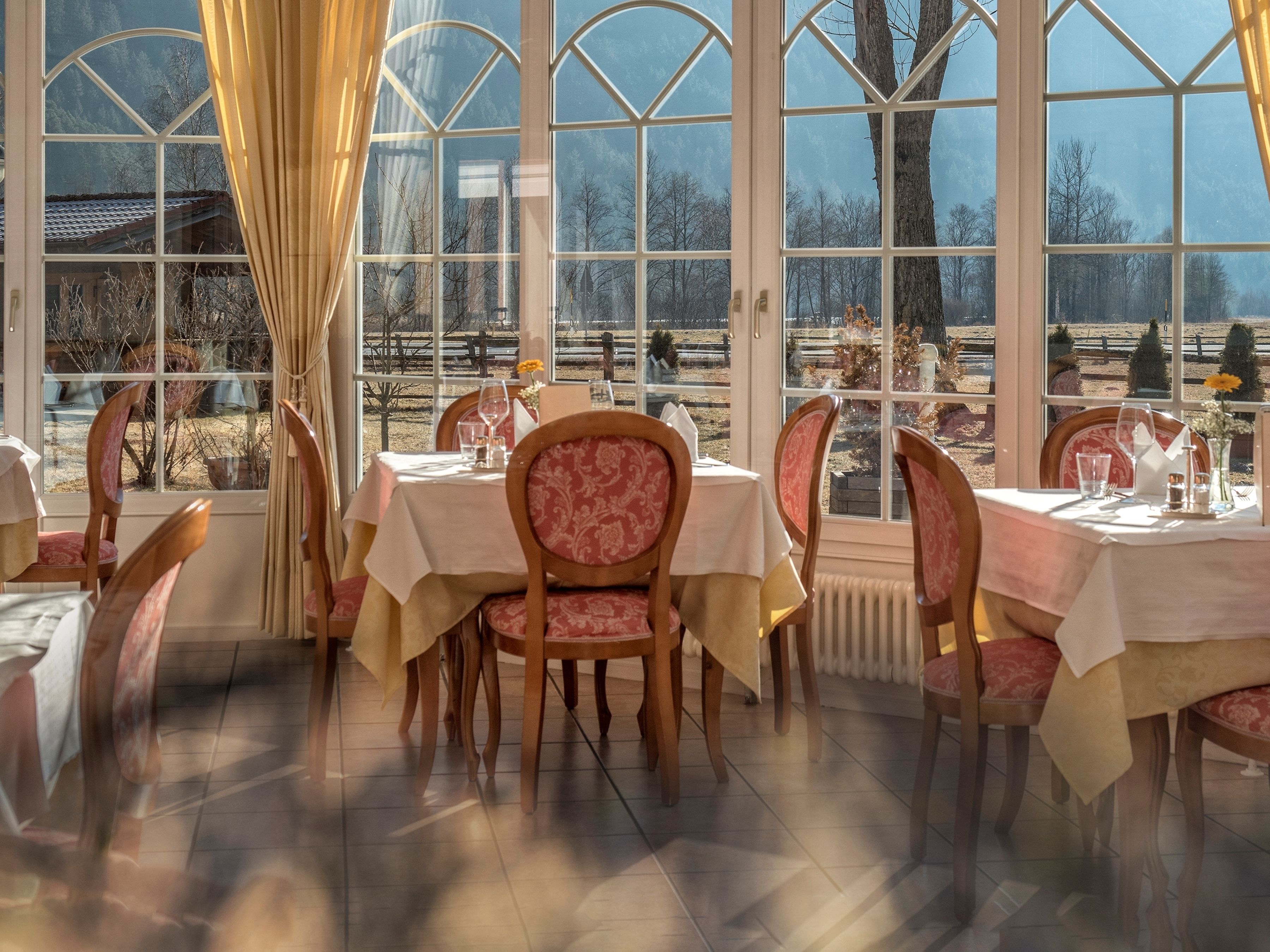 A dining area with tables set for two, large windows, and a view of a park.