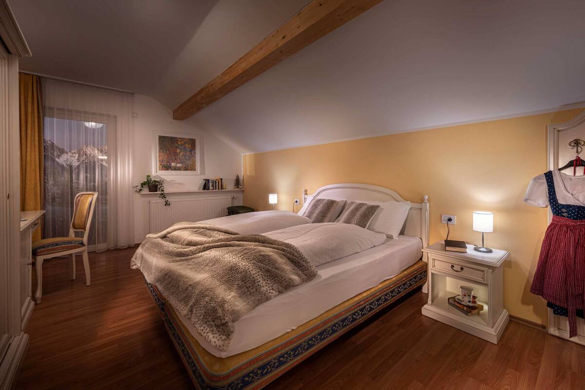 Un dormitorio con una cama grande, una mesita de noche con lámpara, una silla y vistas a montañas a través de una ventana.
