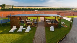 Un espace extérieur aménagé, abrité et doté de chaises longues et de tables, un chemin bordé de briques et une vue sur un terrain de sport en arrière-plan.