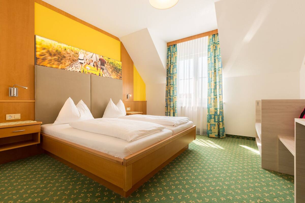 Hotellihuone, jossa on parivuode, työpöytä, tuoli, suuri ikkuna verhoilla ja taulu seinällä.
