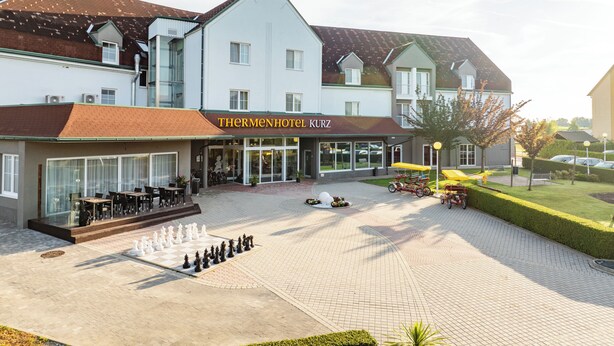 Un hôtel avec un échiquier dans la cour, des places assises en plein air et une enseigne indiquant « Thermenhotel Kurz ».