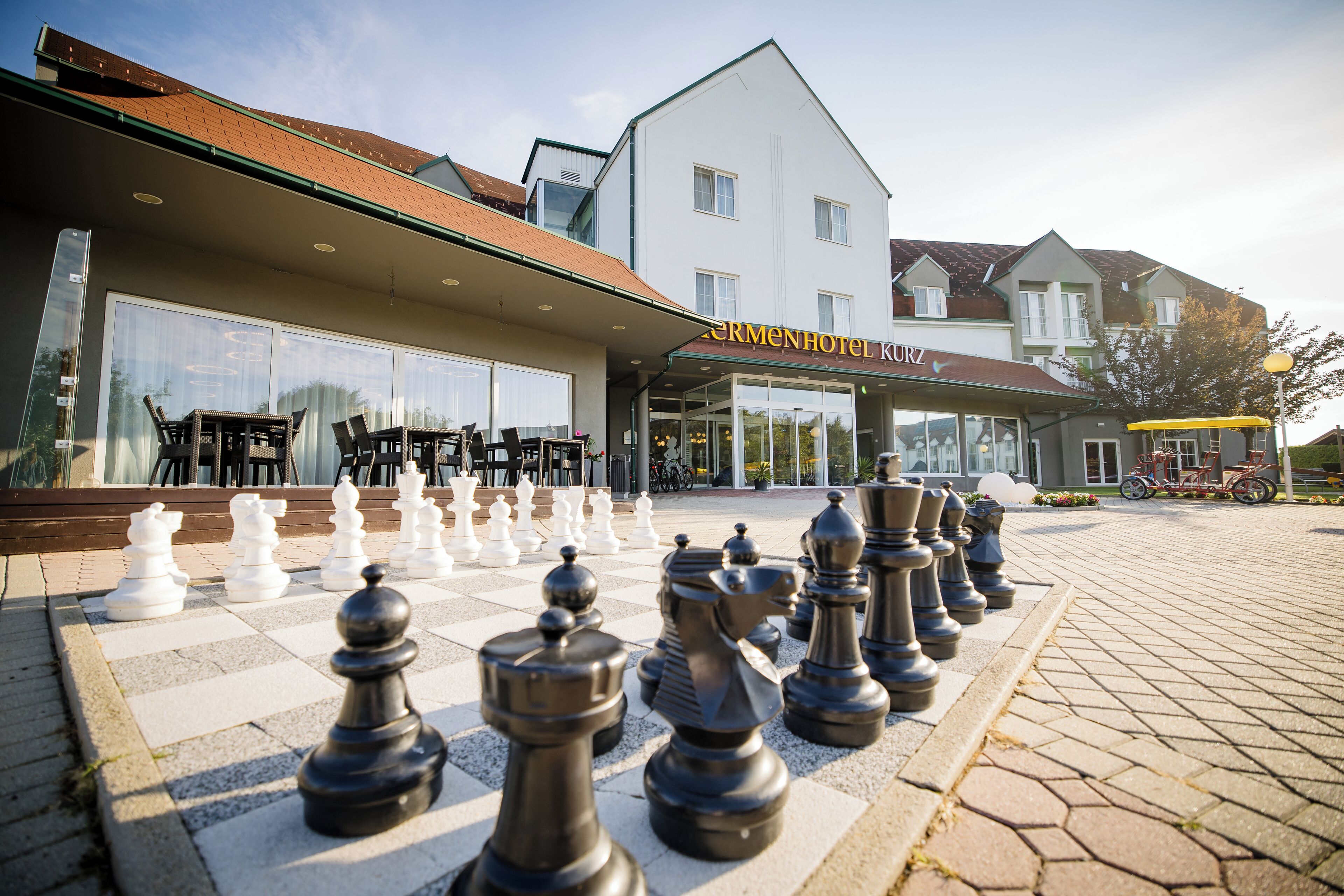 Un jeu d’échecs est installé sur une surface pavée devant un bâtiment sur lequel on peut lire l’inscription « CERMENHOTEL KURZ ».
