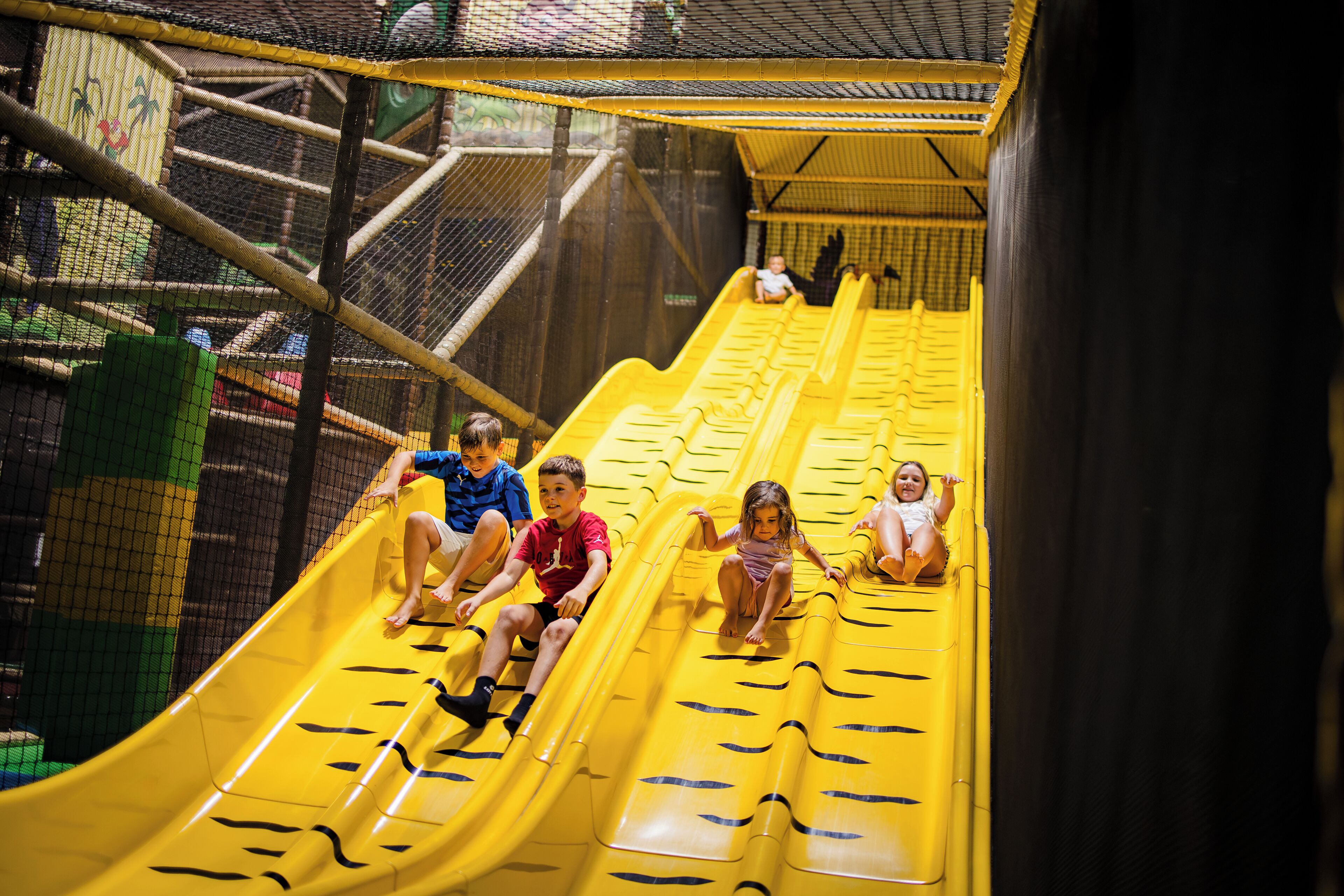 A yellow indoor slide with children at the top, surrounded by safety netting.